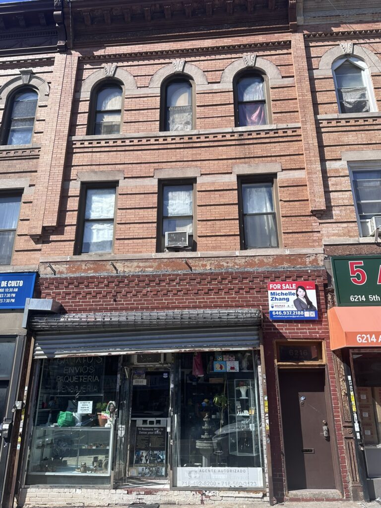 A three-story brick building with arched windows. The ground floor has a storefront with a glass display. A "For Sale" sign featuring a real estate agent is visible next to the entrance. The address number 6216 is displayed on the right.