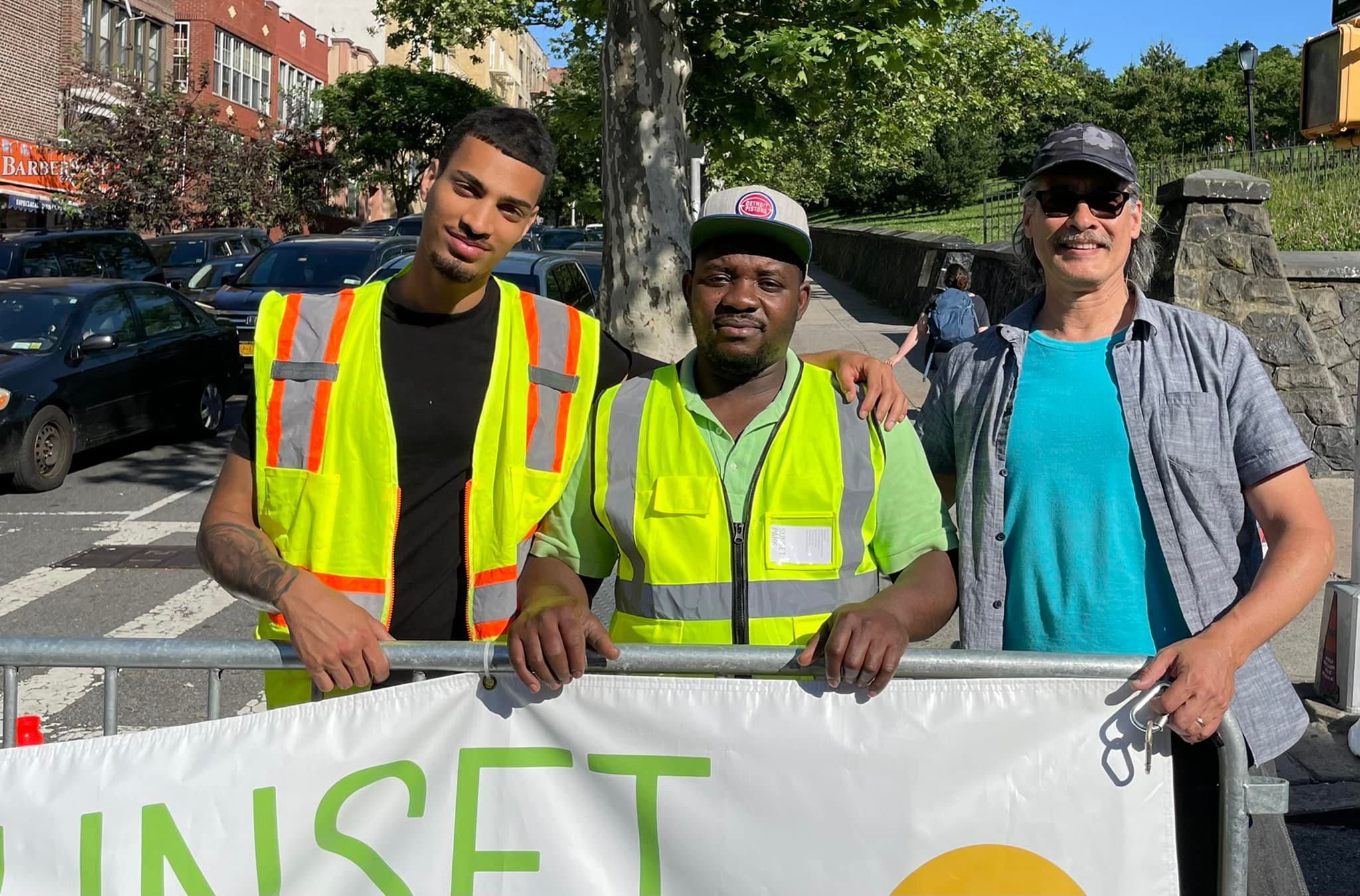 Three men in casual attire, two wearing high-visibility vests, standing outdoors holding a banner with the word "unset" and a circular logo. the background features lush greenery and a clear blue sky.