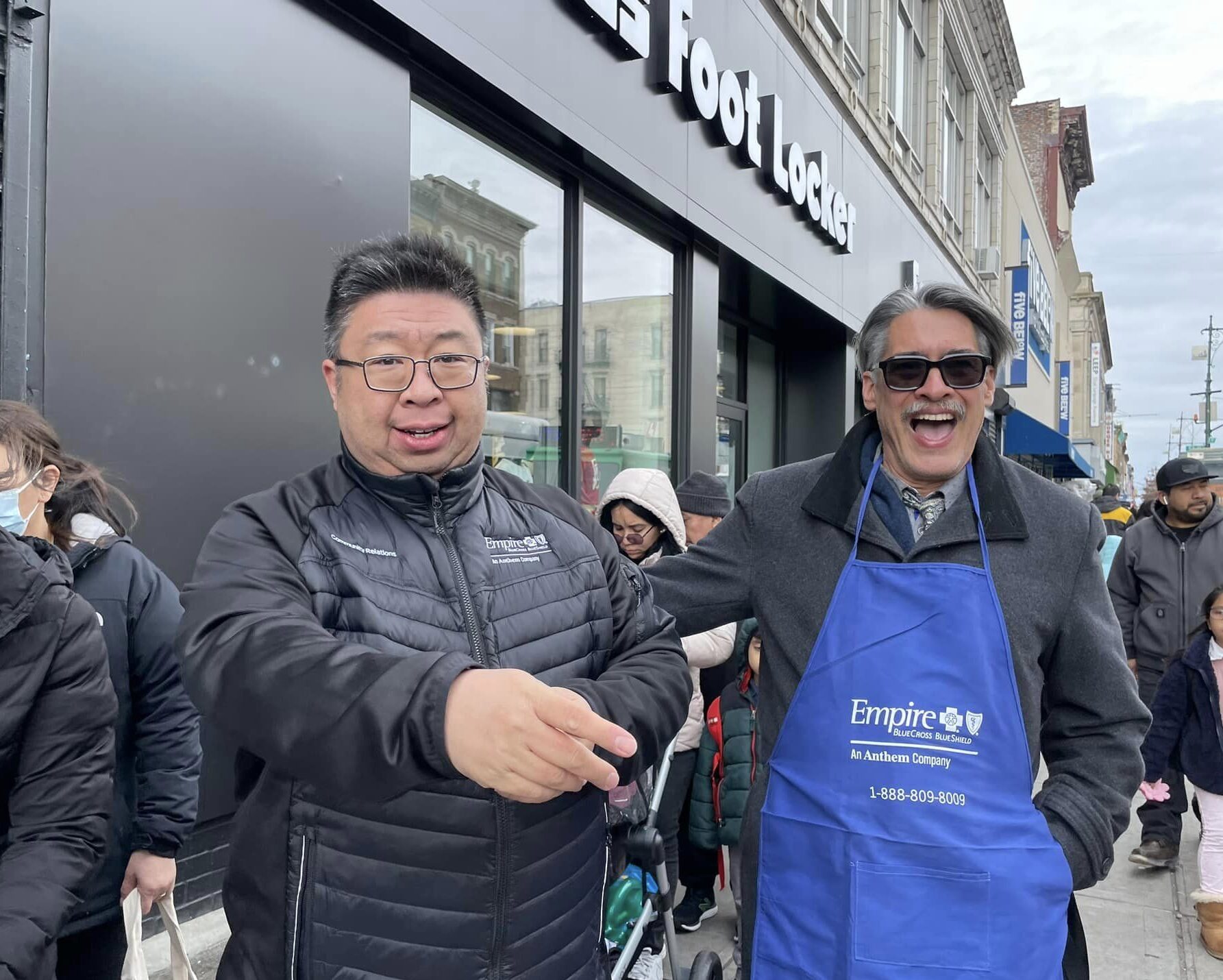 Two men, one wearing a black jacket and one in a blue apron, are smiling and gesturing animatedly on a busy street with people and storefronts in the background.