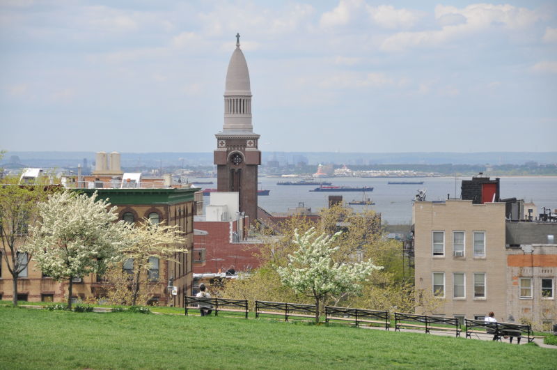 View from a park overlooking a cityscape featuring a tower, blooming trees, and buildings, with a river and distant ships in the background.