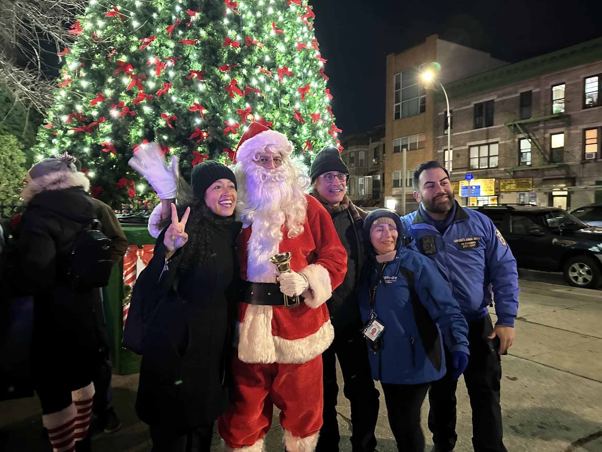 A group of cheerful adults posing with a person dressed as santa claus next to a large, brightly decorated christmas tree outdoors at night.