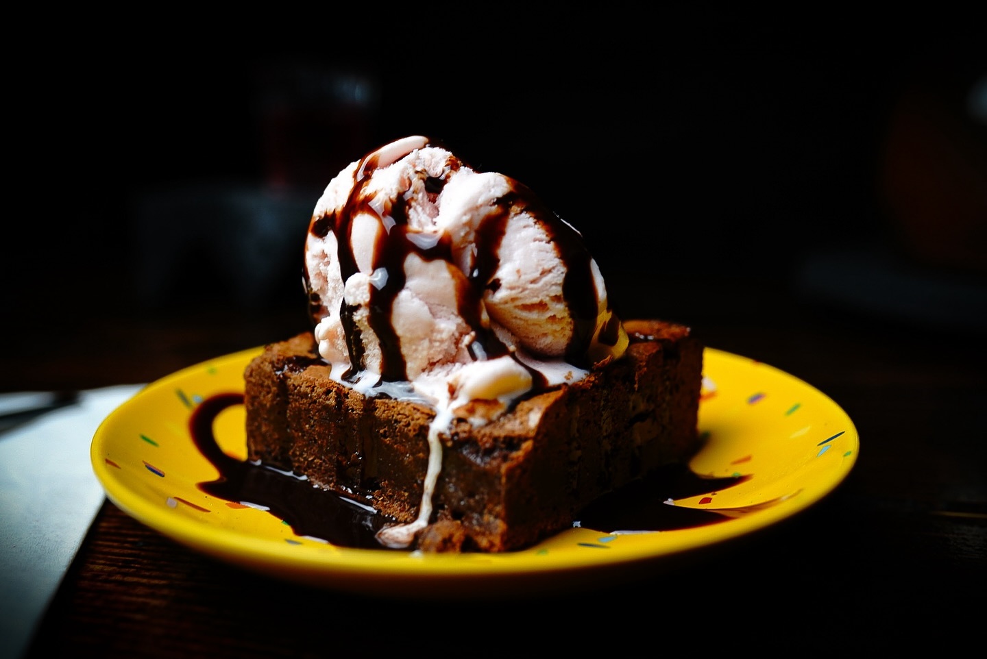 A delectable dessert of vanilla ice cream scoops on a toasted bread piece, generously drizzled with chocolate syrup, served on a vibrant yellow plate.