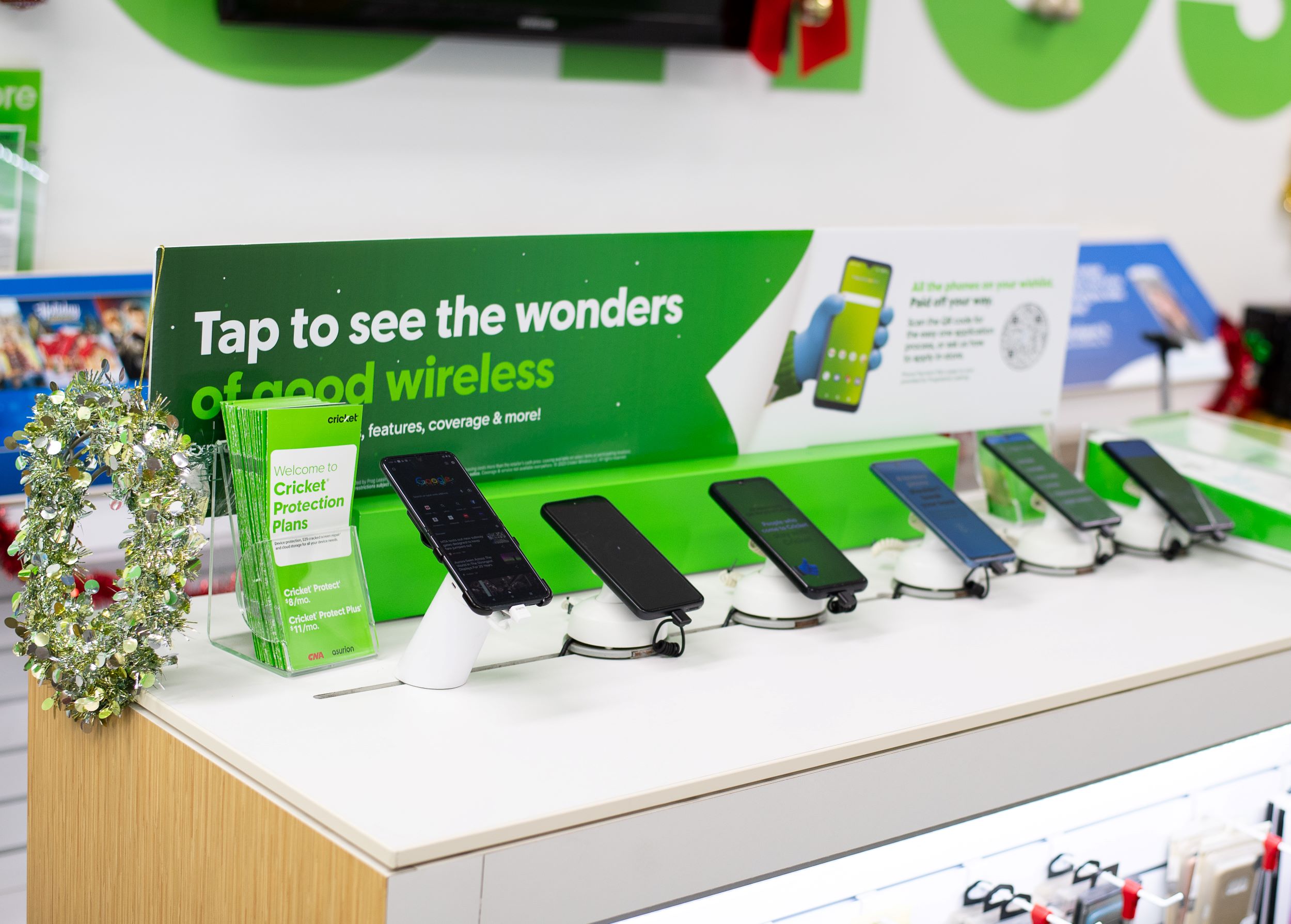 A display of smartphones aligned on a kiosk in a store with promotional signs for cricket wireless, adorned with a small holiday wreath on the side.