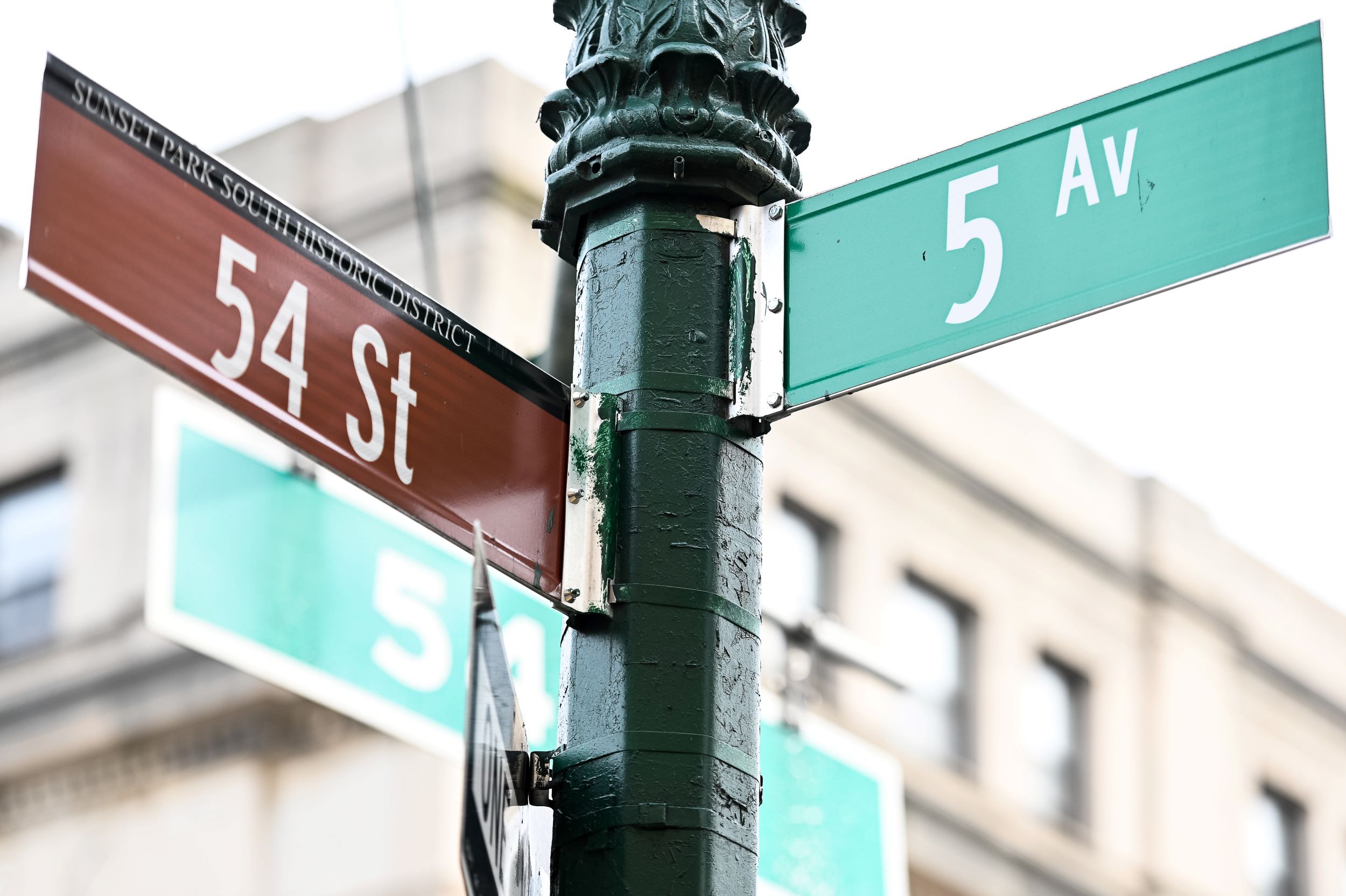 Close-up of street signs at the intersection of 54th street and 5th avenue, mounted on a green pole, with blurred buildings in the background.