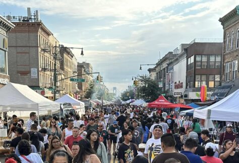 A bustling street festival with many people walking among vendor tents under a clear sky, flanked by city buildings on both sides.