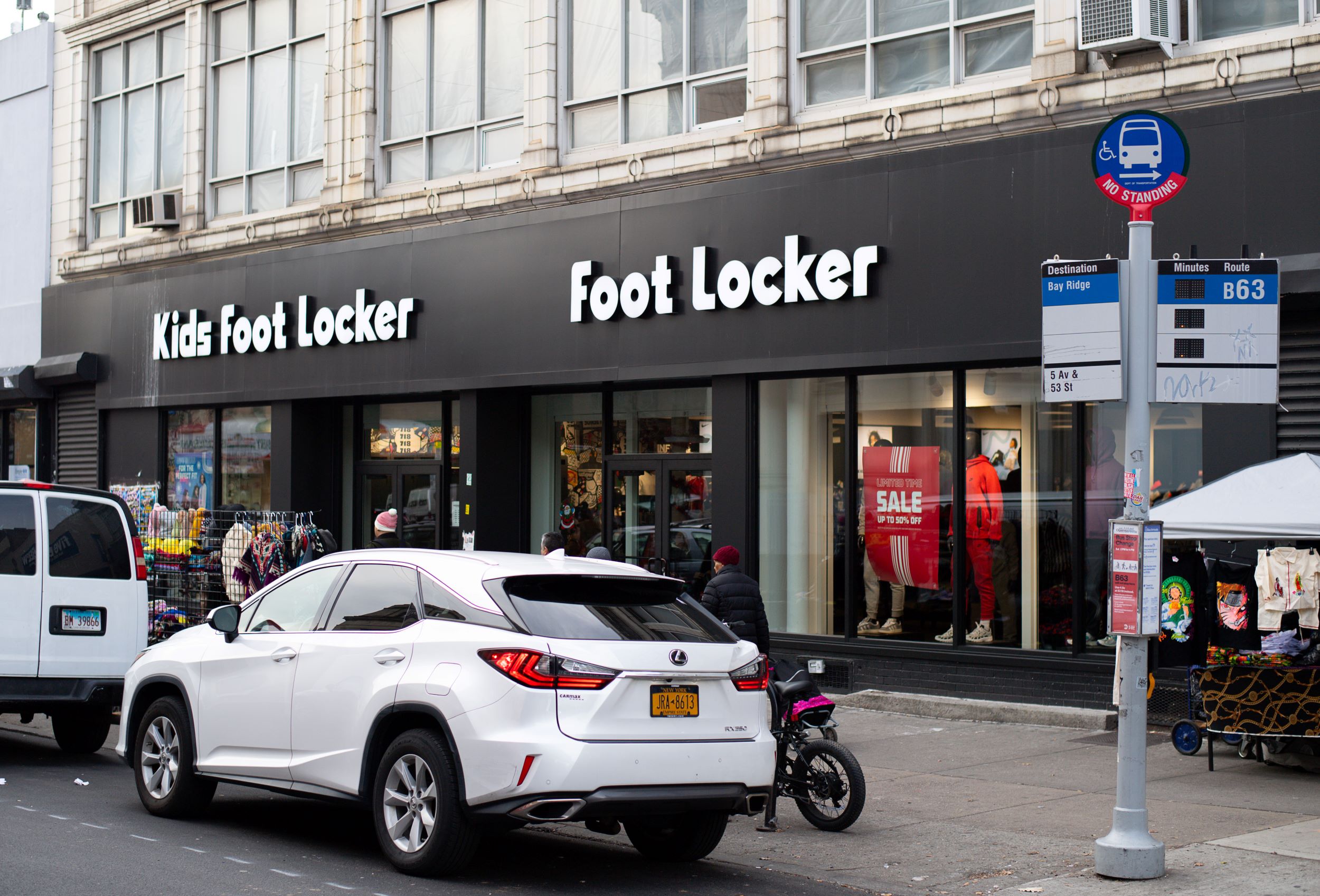 Street view showcasing a foot locker store with visible signage, adjacent to a kids foot locker, with cars and a bus stop sign in the foreground.