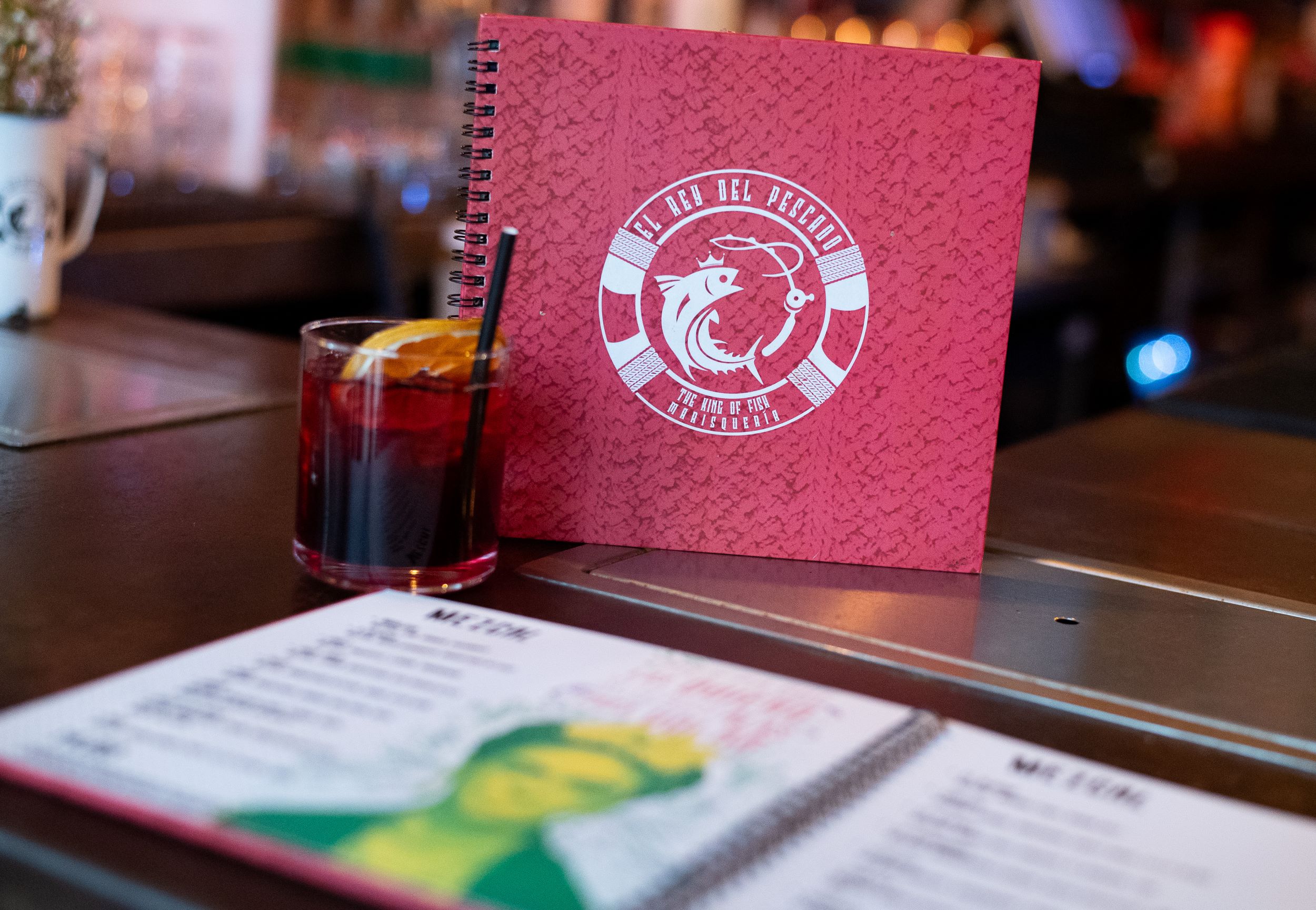 A menu from "le bar belge" standing on a bar table with a pink textured cover featuring a white emblem, next to a dark beverage in a small glass.