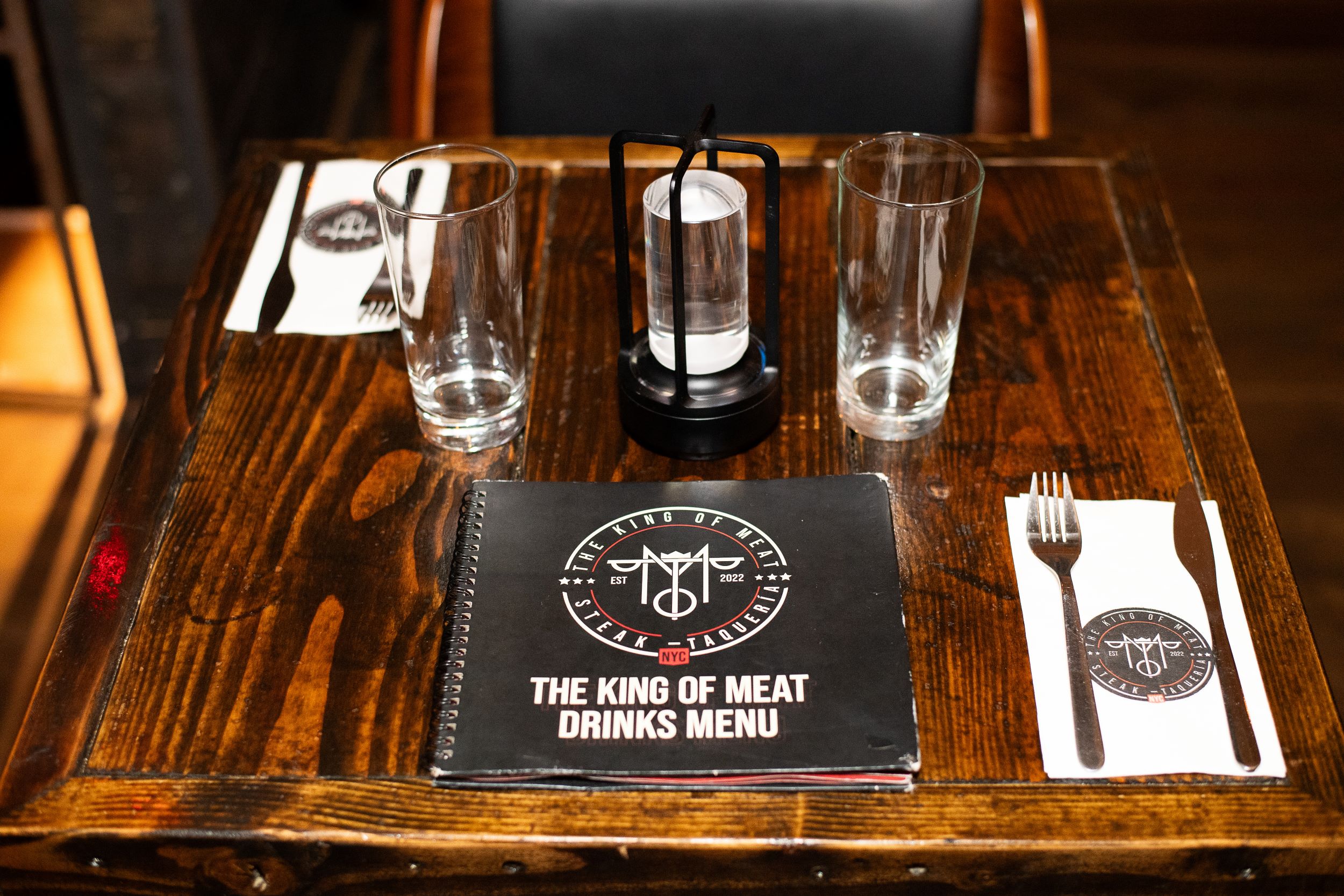 A wooden table set for two at a restaurant, featuring a drinks menu titled "the king of meat," two glasses, a lantern, and cutlery wrapped in napkins with a logo design.
