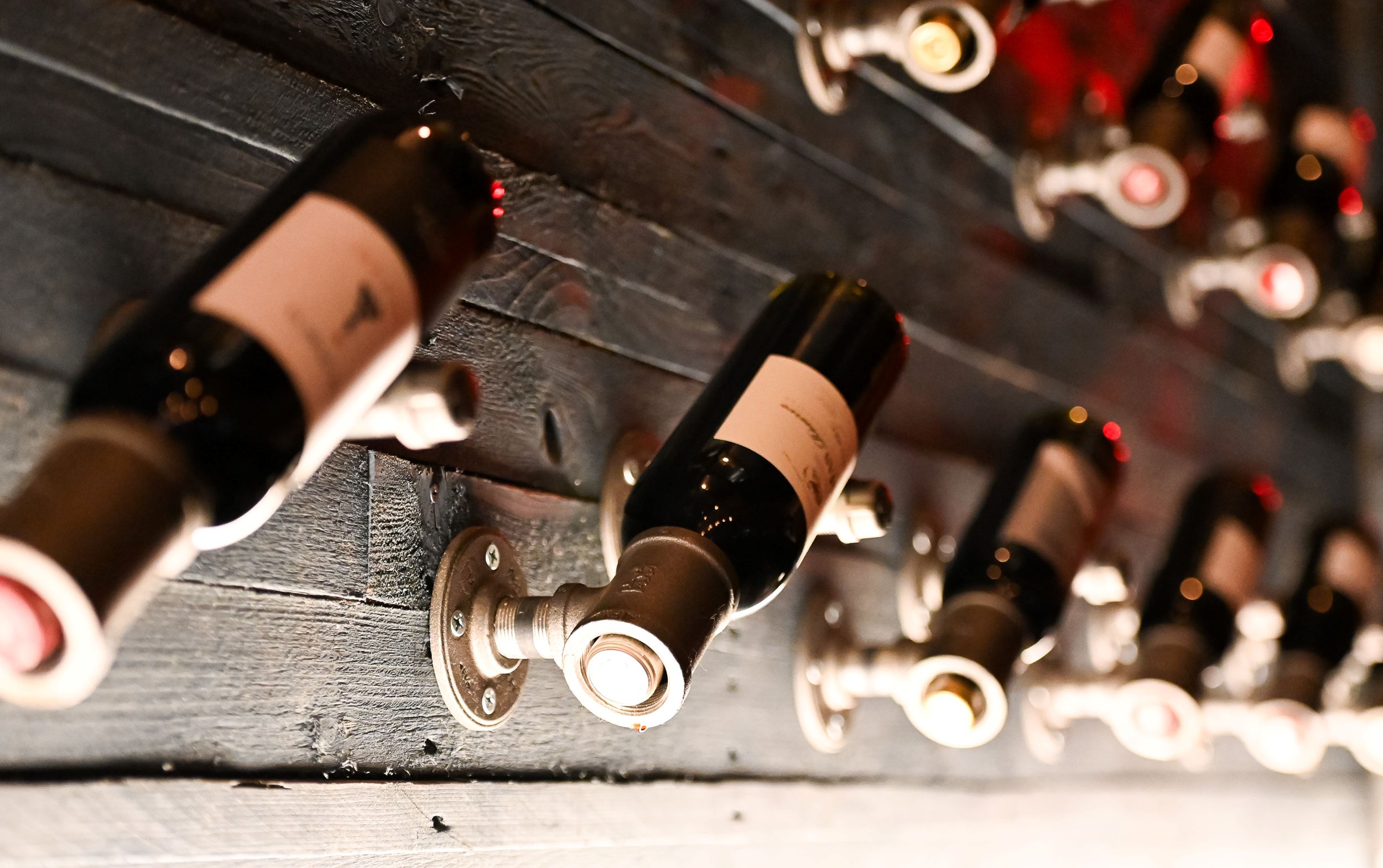 A display of wine bottles mounted diagonally in metal holders against a dark wooden wall, viewed from a low angle with a focus on the closest bottle.