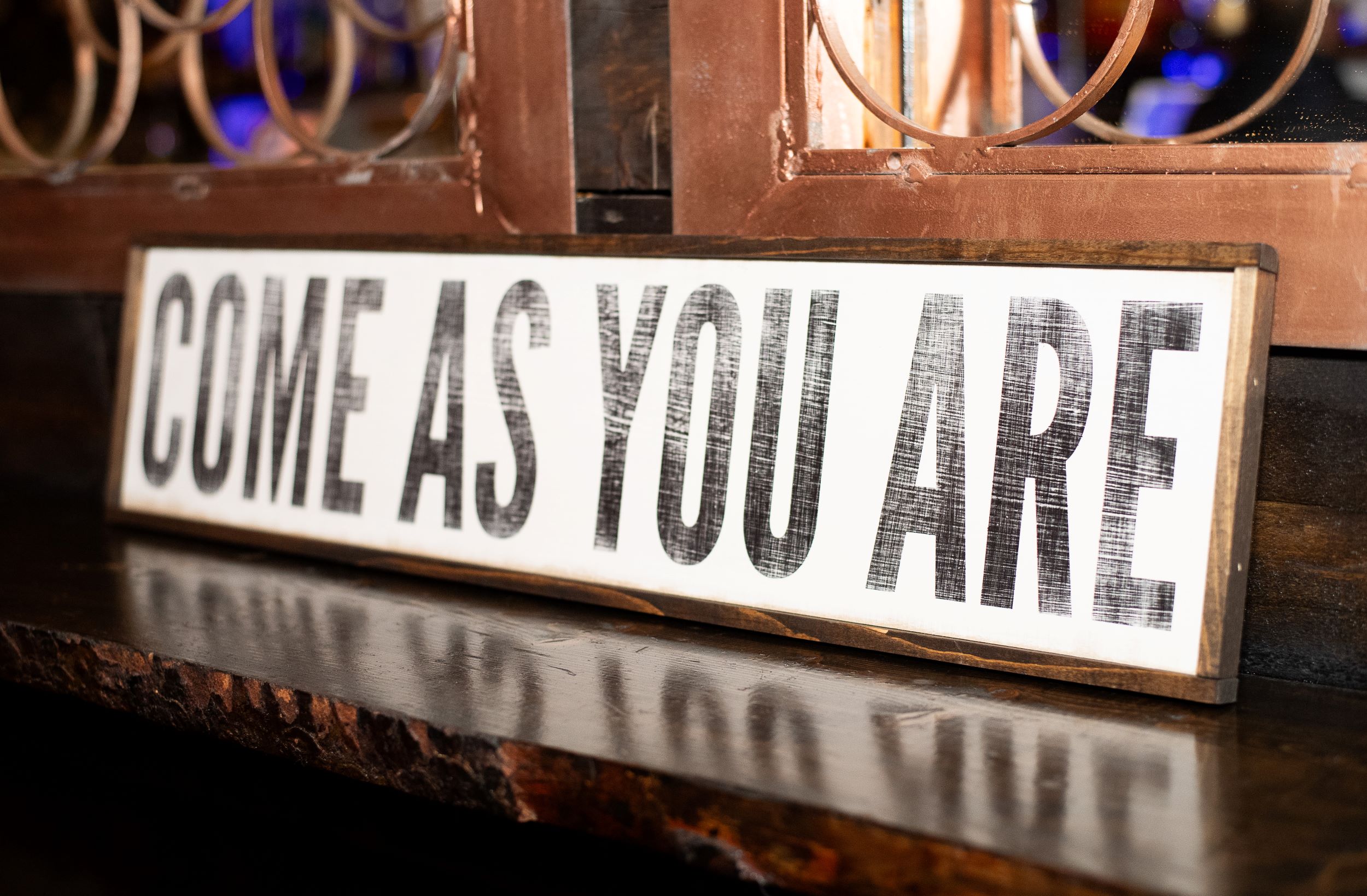 Sign that reads "come as you are" in bold, black letters on a white background, displayed on a wooden counter with blurred lights in the background.
