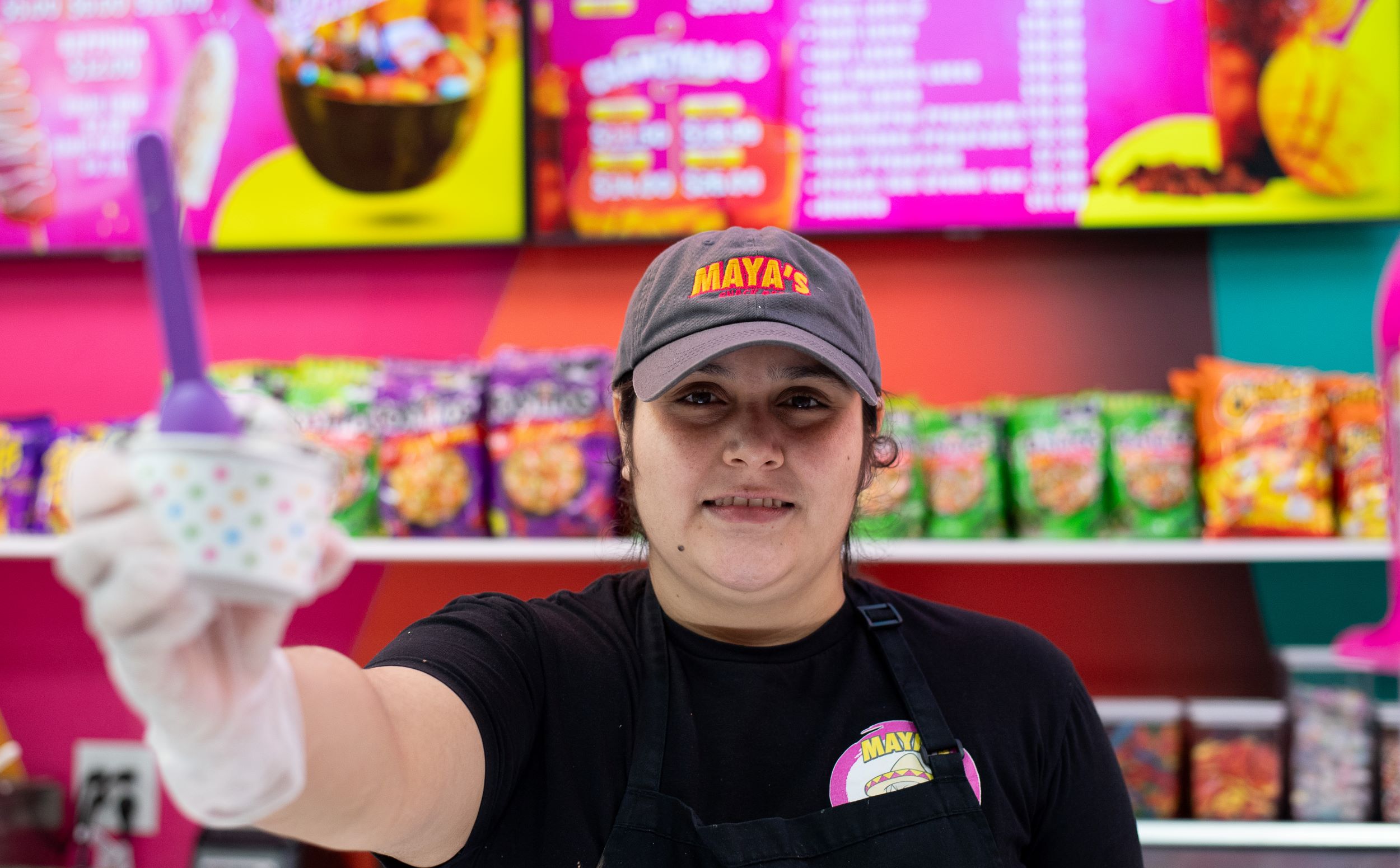 A woman in a black apron and cap labeled "maya's" holds out a cup of frozen yogurt towards the camera, standing at a brightly colored food counter.