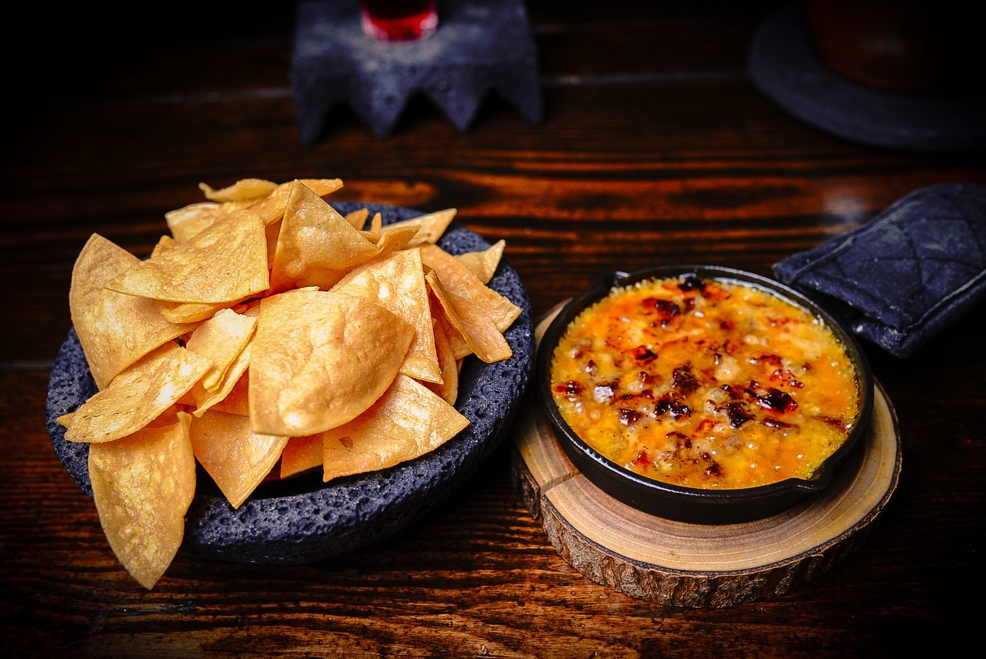 A bowl of crispy tortilla chips beside a dish of bubbling hot cheese dip, served on a wooden table with rustic decor.