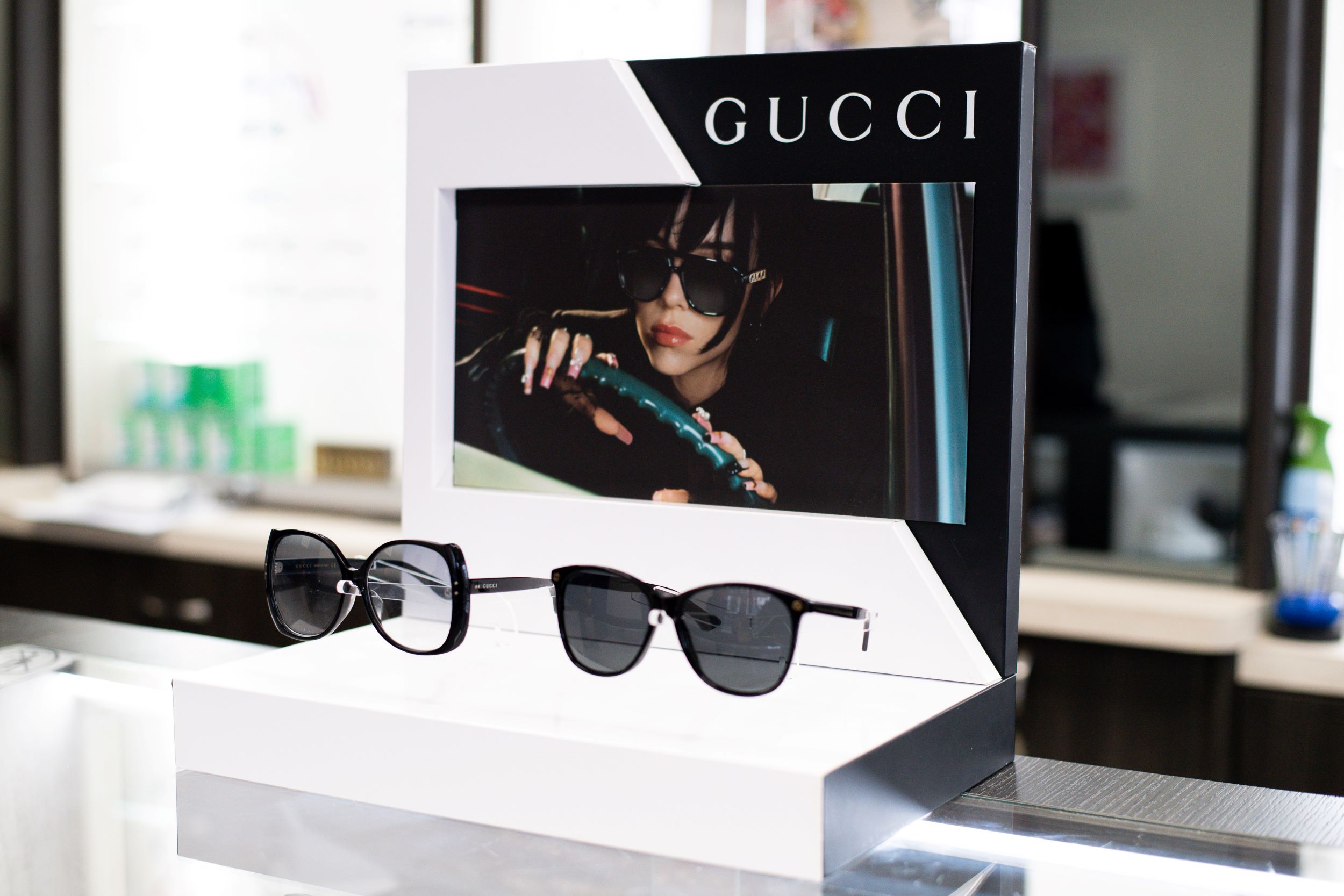 A display stand for gucci sunglasses featuring a black and white poster with the brand name and a photo of a woman wearing sunglasses. a pair of sunglasses rests in front on a white platform.