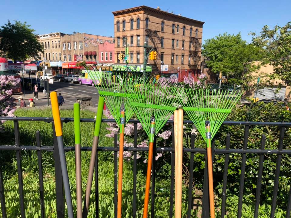 Row of green garden rakes leaning on a metal fence with a colorful urban backdrop, featuring low-rise buildings and a clear blue sky.