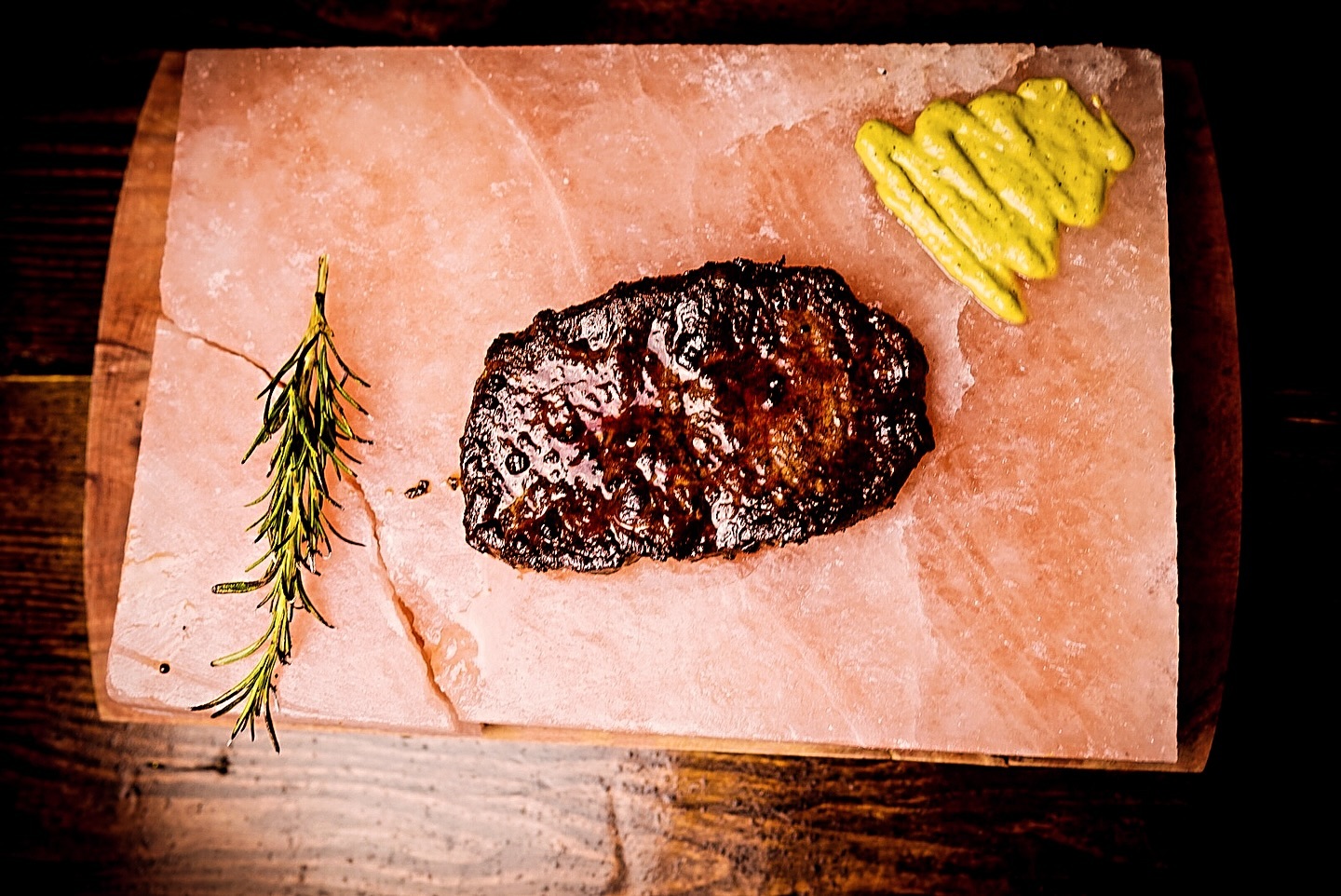 Grilled steak on a pink salt block with a sprig of rosemary and a dollop of mustard on the side. the dish is presented on a dark wooden table.