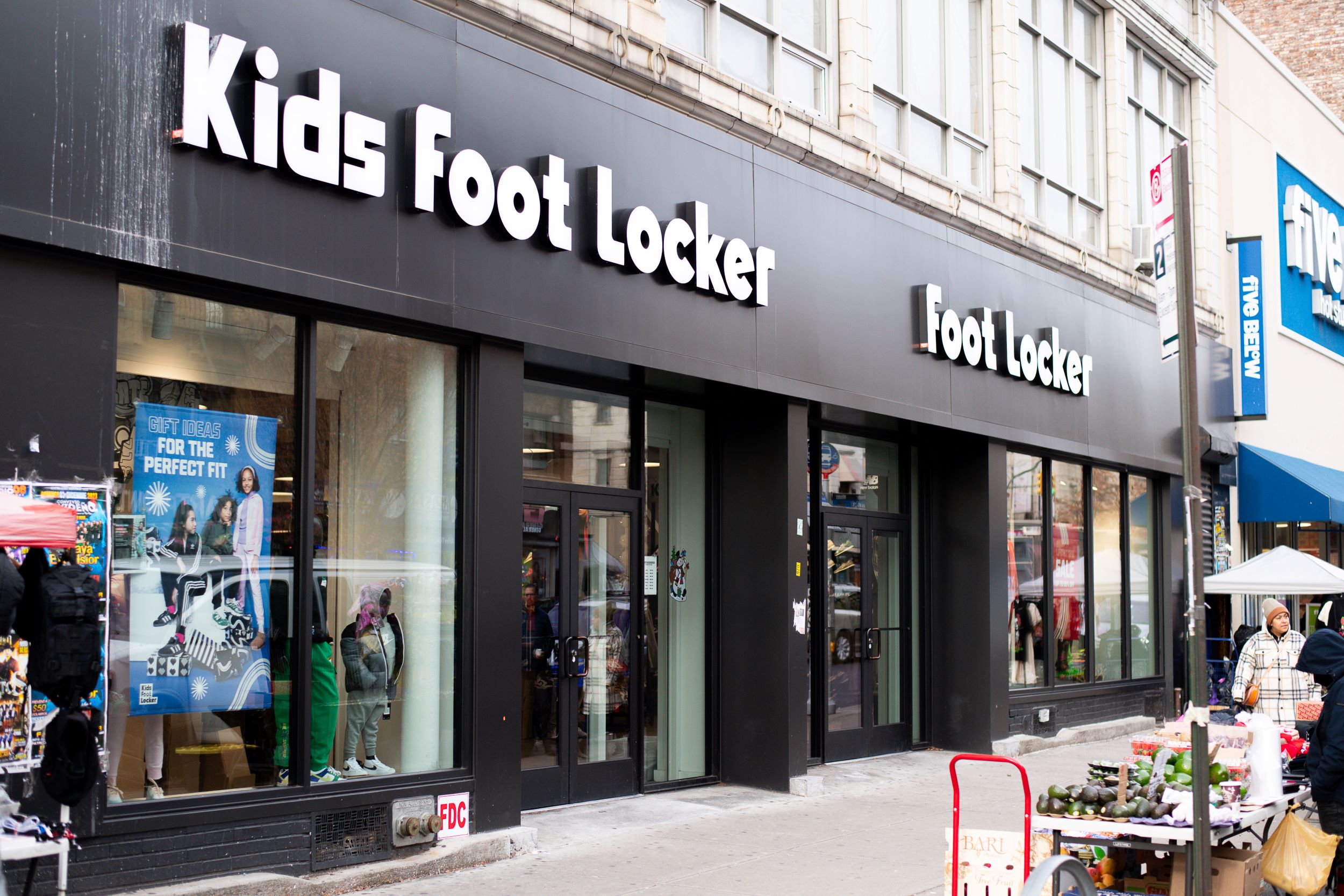 Exterior of a kids foot locker store with displayed sneakers and apparel visible through large glass windows, situated on a busy urban street.