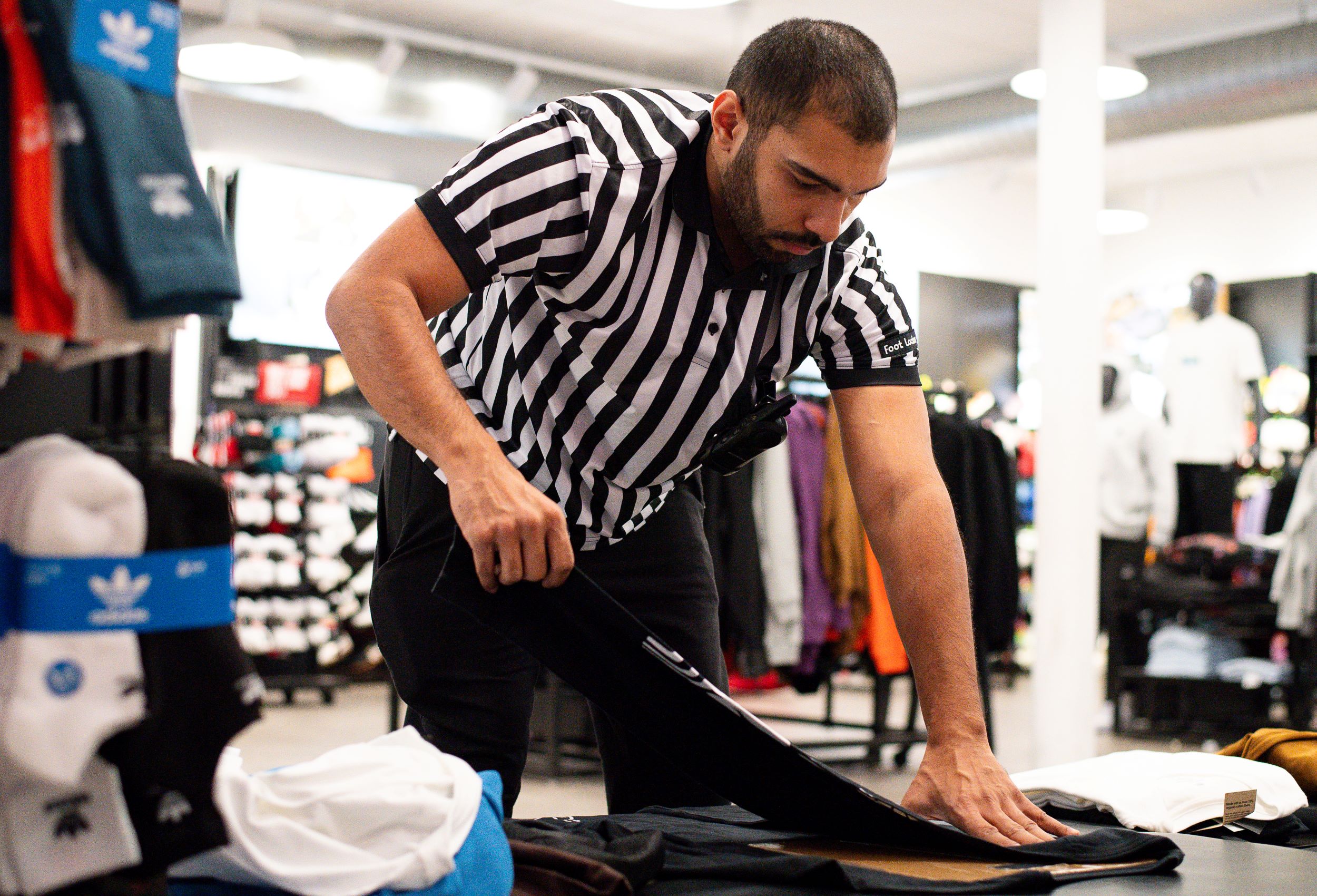 A man in a striped referee shirt folding clothes in a retail store, surrounded by various apparel items.
