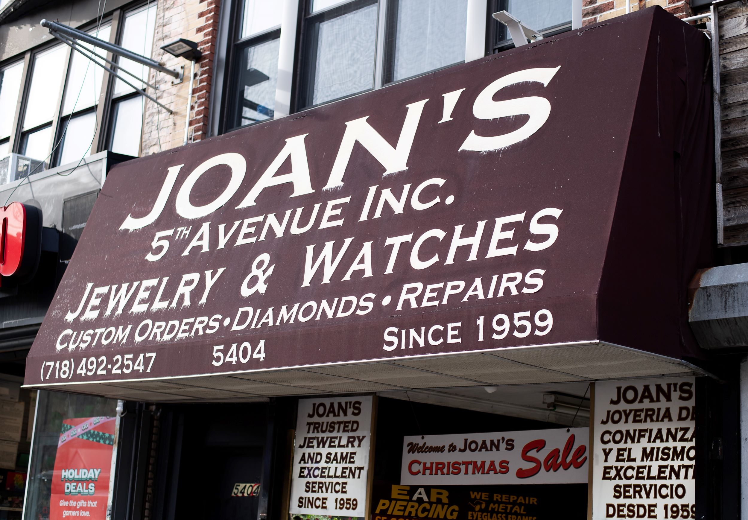 A weathered outdoor sign for "joan's 5th avenue inc., jewelry & watches" indicating services like custom orders, diamonds, and repairs, with the establishment date "since 1959." a faded sale banner is visible beneath.