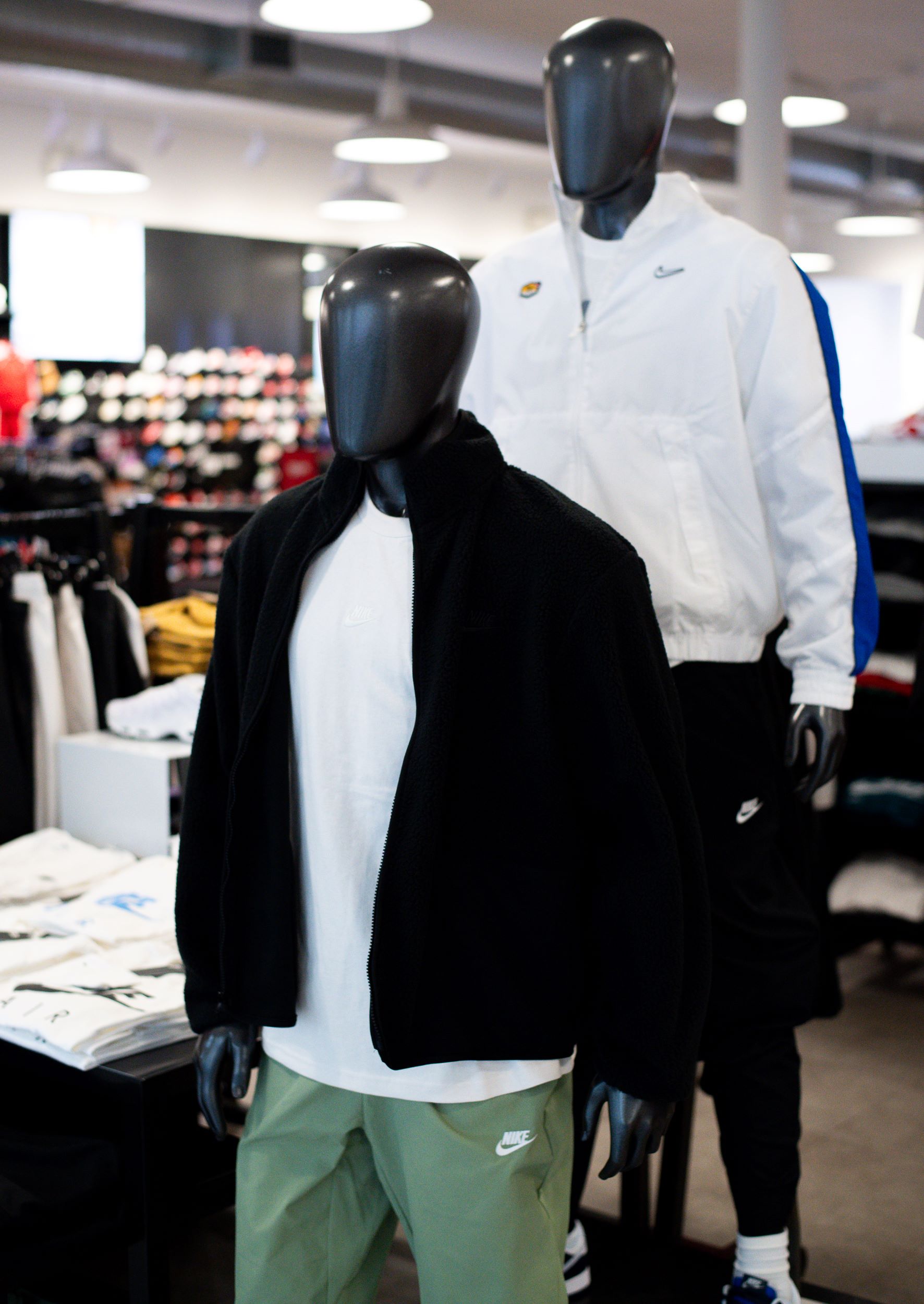 Two mannequins dressed in nike apparel standing inside a store, one wearing a black hoodie and green shorts, the other in a white and blue jacket.