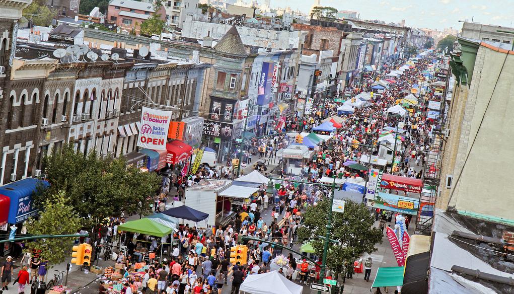 Aerial view of a bustling street festival in an urban area, showing dense crowds, vendor stalls, and historic facades along a tree-lined avenue.