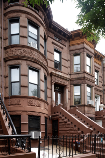 A Brownstone Building In An Urban Area, Featuring Curved Bay Windows, Ornate Stonework, And A Staircase Leading To A Black Entrance Gate.