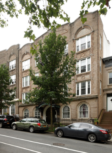 A Tree-lined Street Featuring A Row Of Brick Apartment Buildings With Arched Windows And Entryways, Parked Cars Along The Curb.