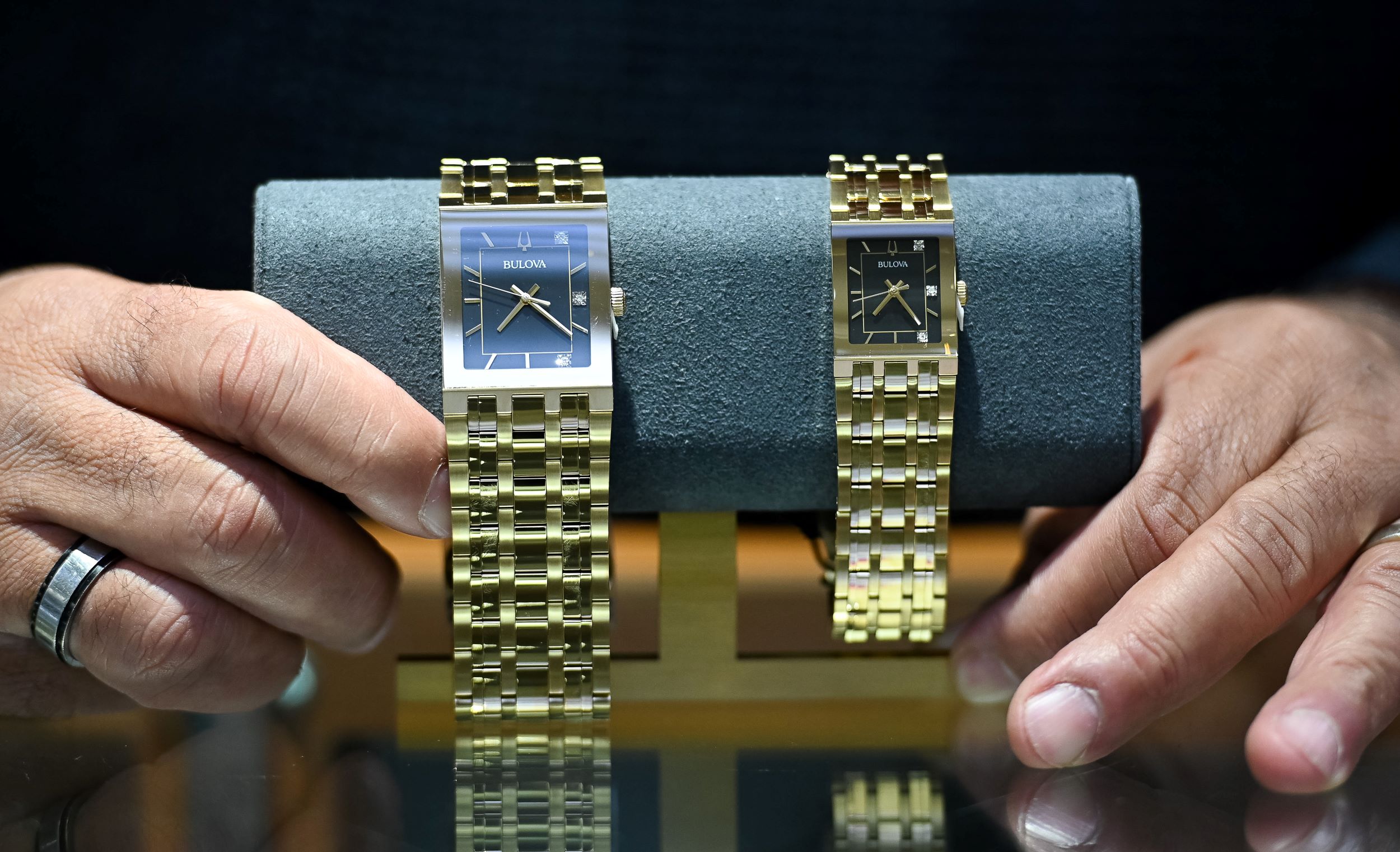 A person displaying two luxury gold watches placed on a gray cushion, with a focus on their hands and the watches' details.