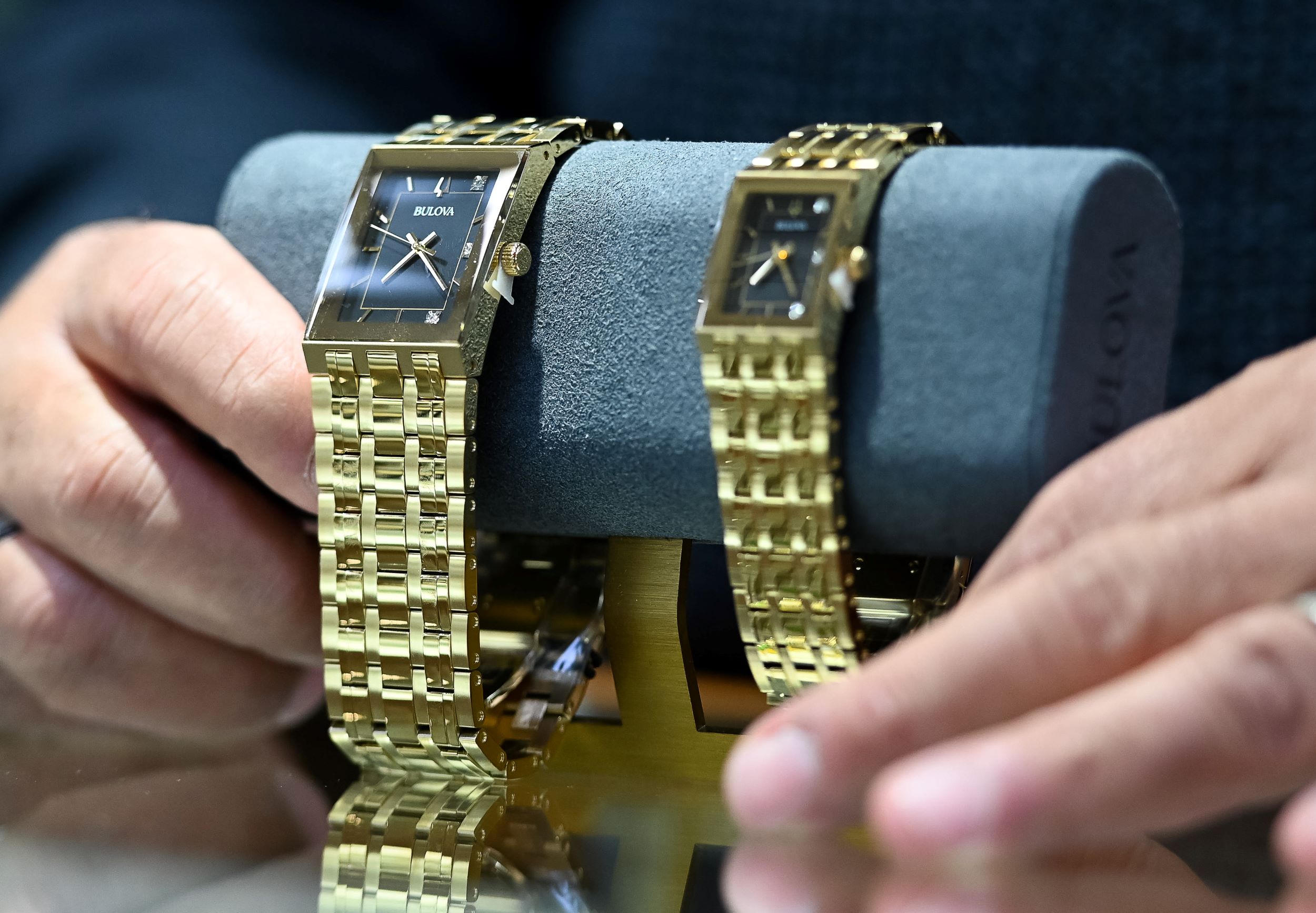 A person's hands adjusting two luxurious gold bulova watches displayed on a gray cushion, highlighting the elegant design and reflective surfaces.