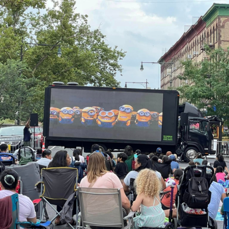 Outdoor movie screening showing minions on a large mobile screen with a crowd of viewers seated on chairs and on the ground in an urban park setting.