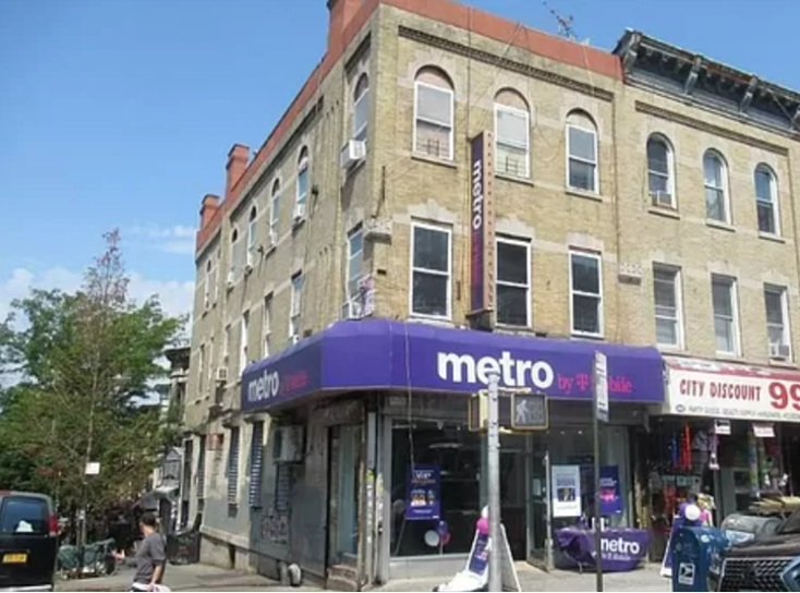 A street corner with a two-story building housing a metro by t-mobile store on the ground floor. the store has purple branding and sits beneath blue skies on a sunny day.