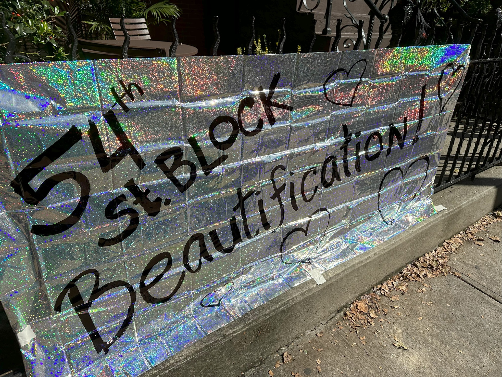 A glittering sign reads "54th st. block beautification" in bold, black letters, displayed in front of a fence on a sunny day.