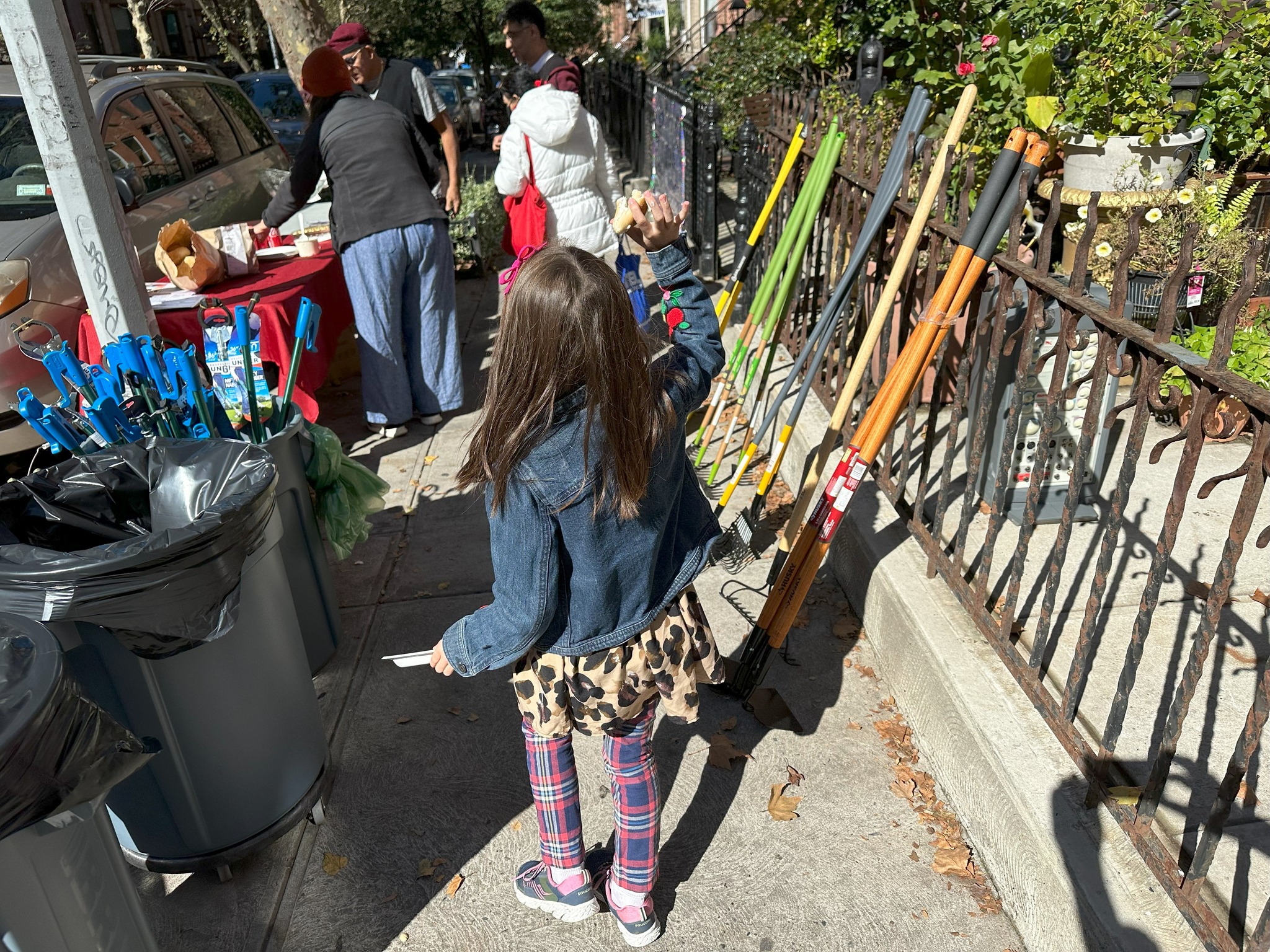 A young girl in a denim jacket and patterned leggings stands on a sidewalk, looking at a rack of colorful brooms for sale at a street market.