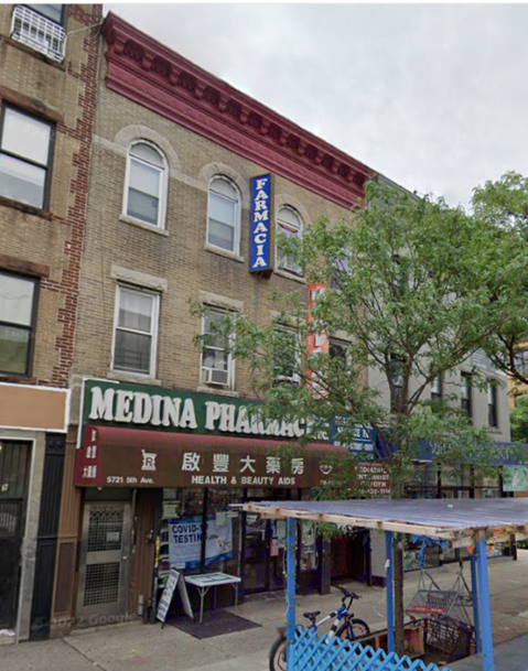 A street view of a three-story brick building with "medina pharmacy" on the ground floor, featuring a blue awning and signs for health and beauty aids. a blue bicycle and trees are visible in front.