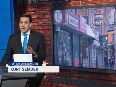 A news anchor in a blue suit presents a story with a backdrop image showing a city street with various storefronts, including a bakery and café. his name and twitter handle appear on the screen.
