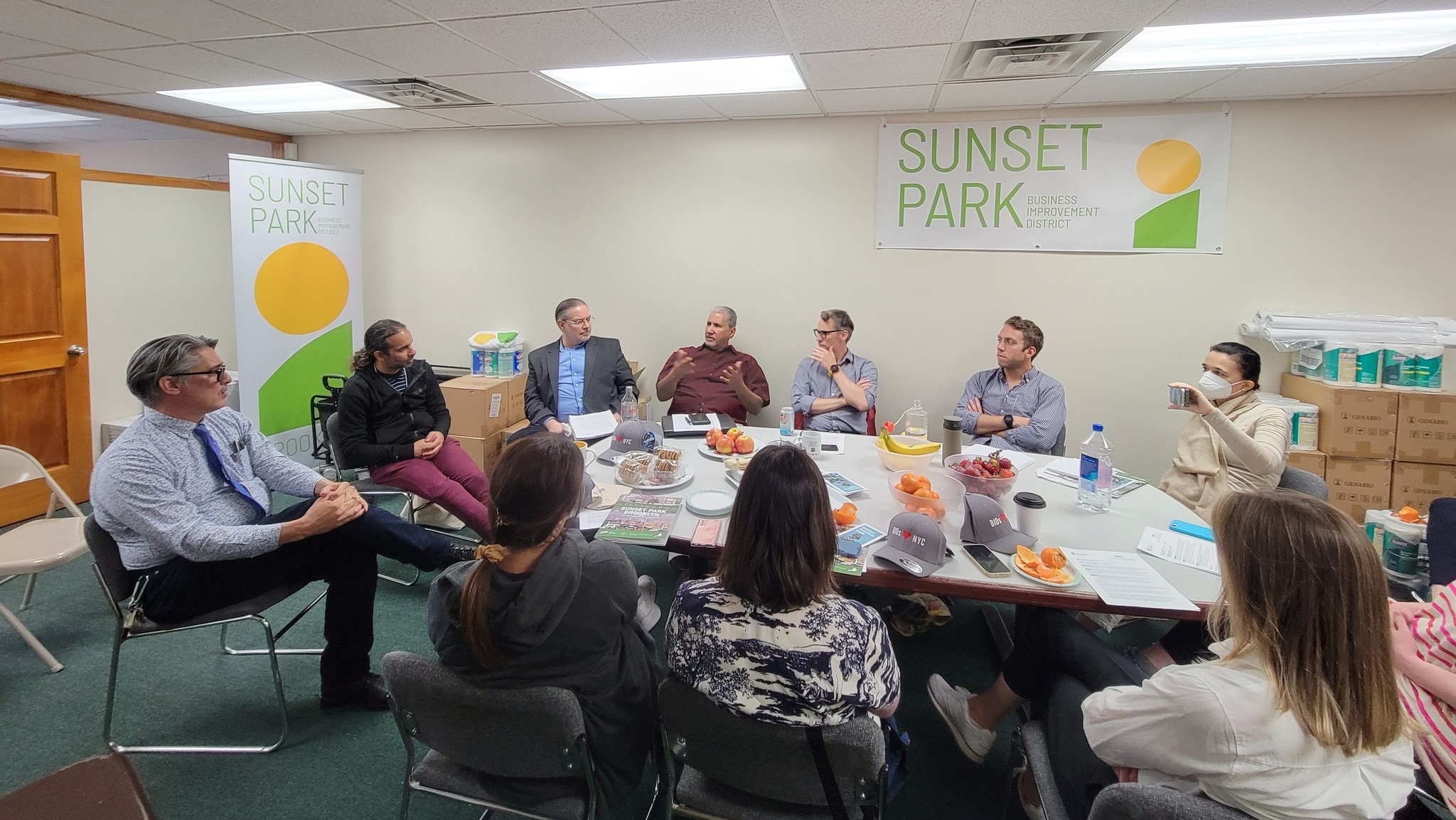 A diverse group of professionals is engaged in a meeting around a conference table, discussing issues, in a room with "sunset park" banners on the walls.