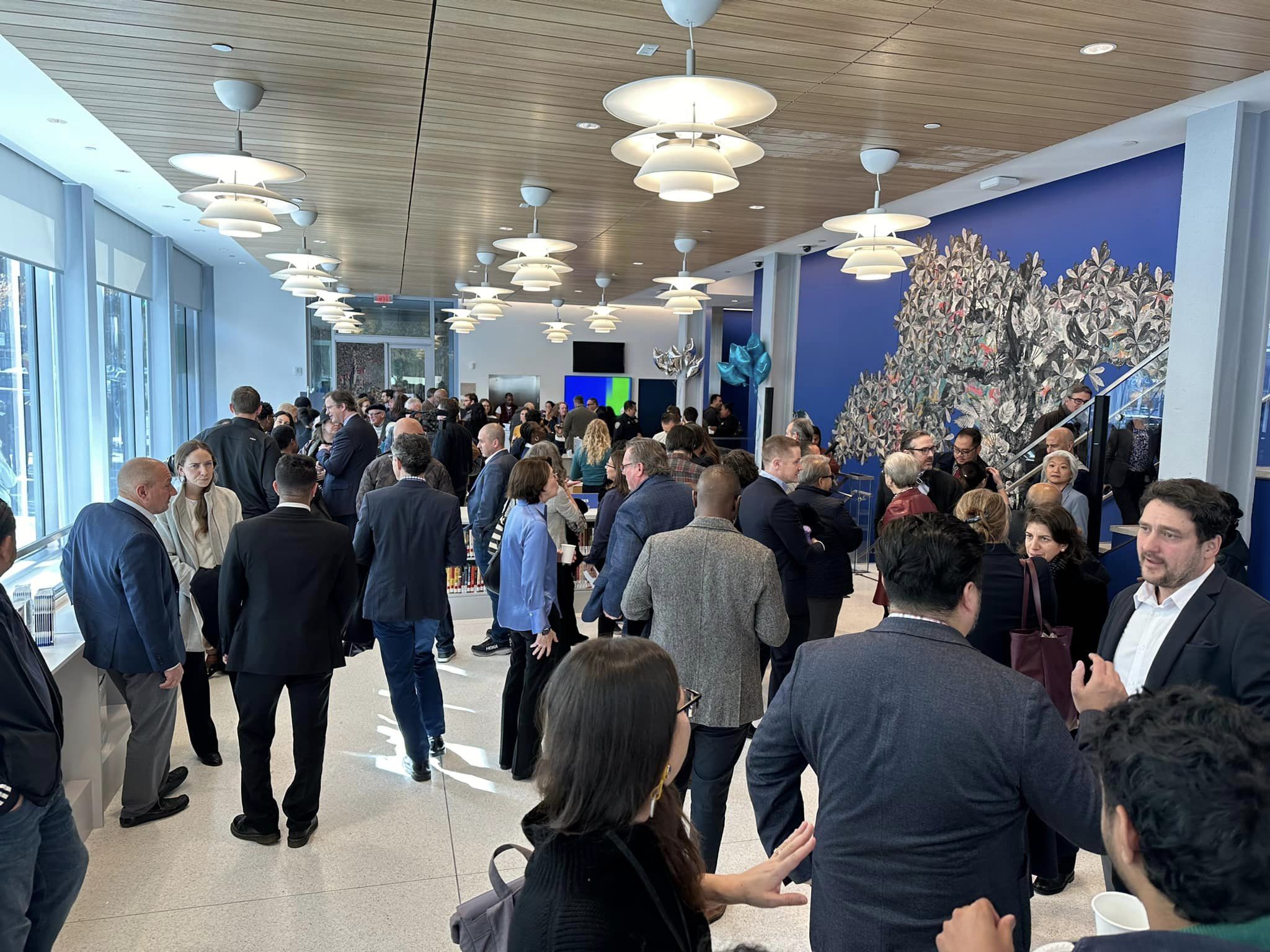 A busy indoor networking event with a crowd of people in business attire talking and mingling in a modern hall with blue walls and spherical light fixtures.