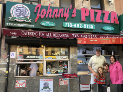 A family of four stands in front of johnny's pizza, a local pizzeria with a red storefront and images of pizza on the windows. there appears to be a worker inside the shop.