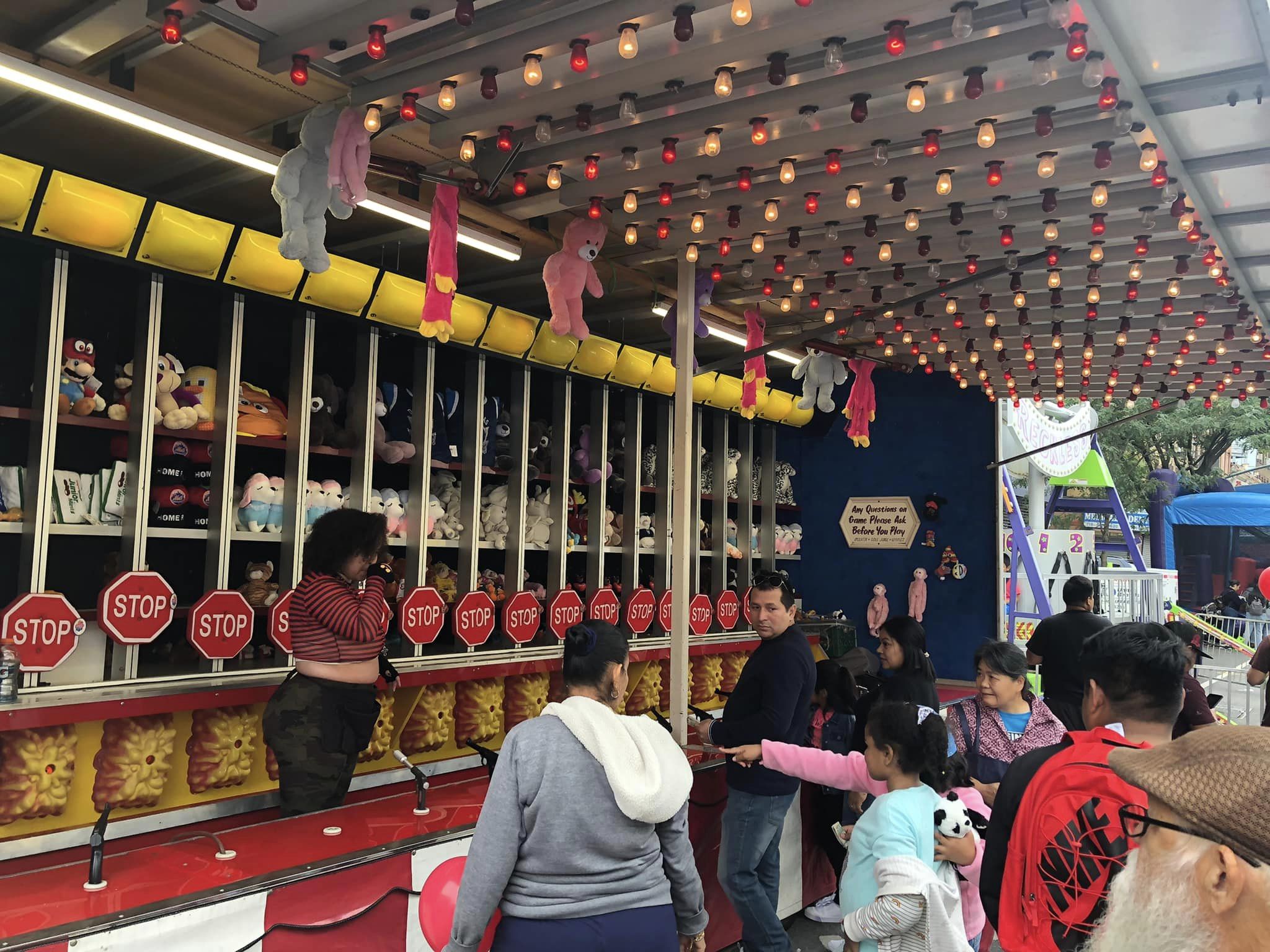 A bustling carnival game booth with people playing hit-a-mole, decorated with lights and plush prizes on display. a child points excitedly, and adults accompany their kids around the booth.