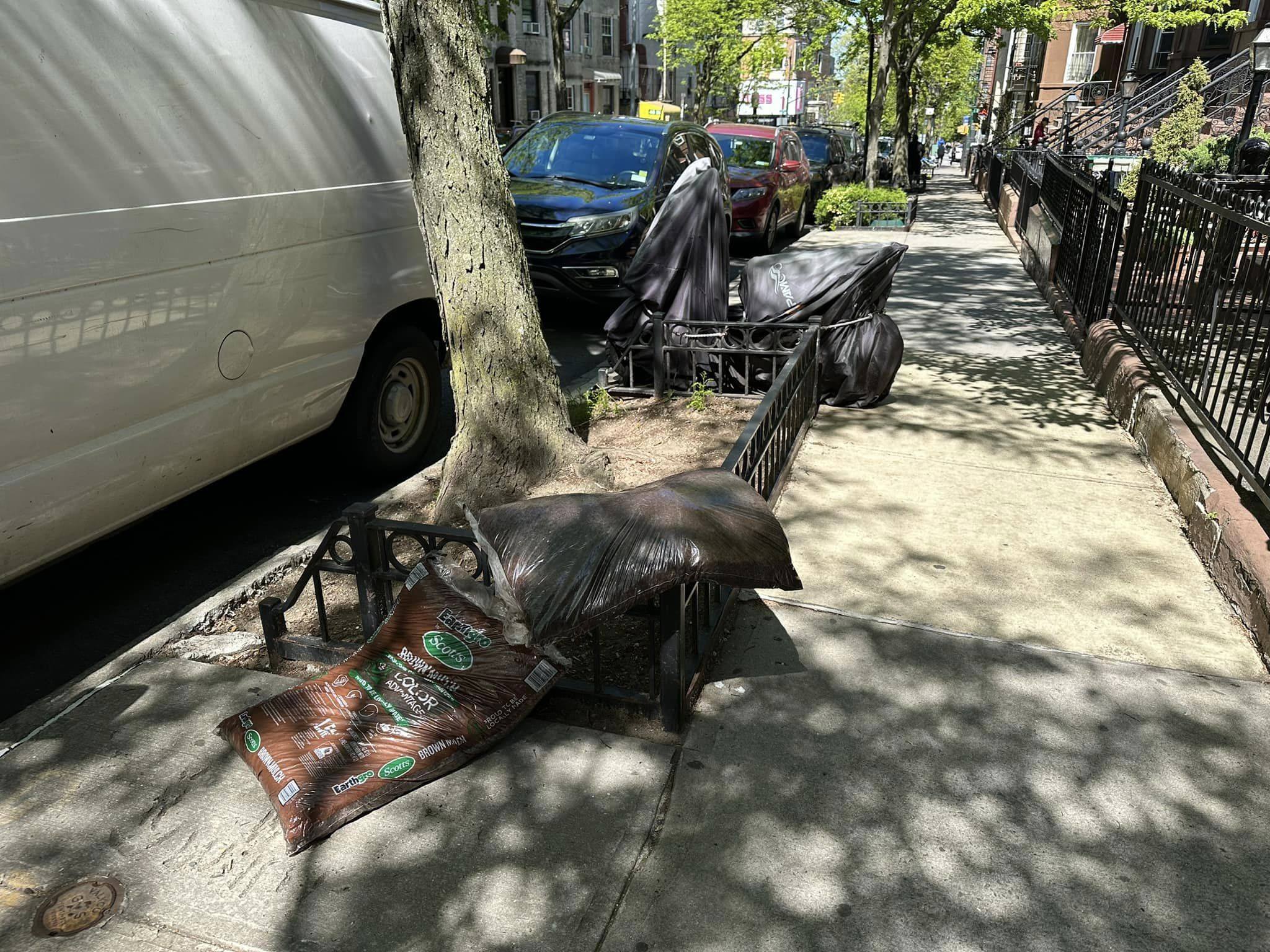 A city sidewalk lined with trees, with a small pile of garbage bags and a ripped open bag of soil near a parked van and covered motorbikes on a sunny day.