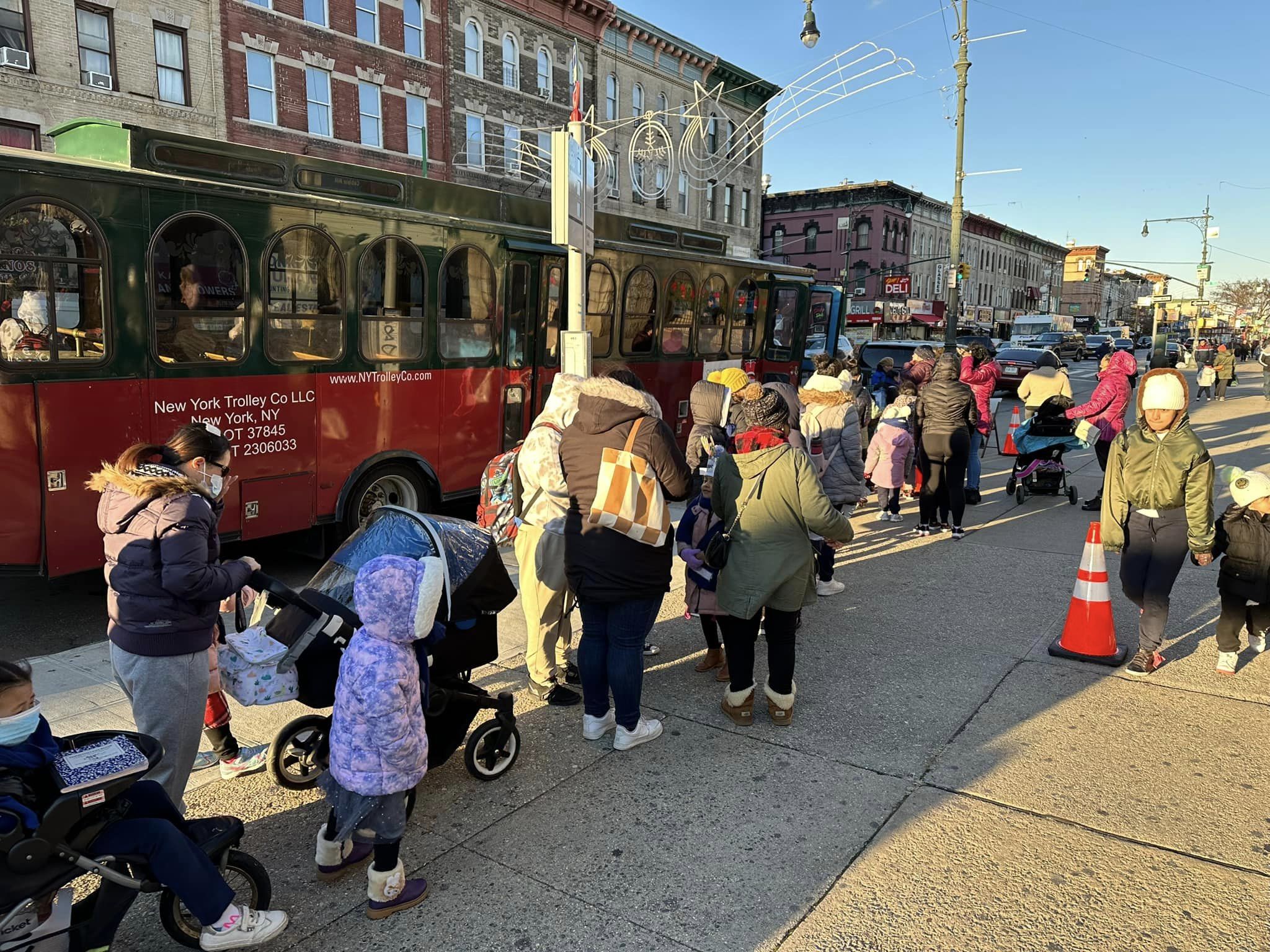 People boarding a red new york trolley co. bus on a sunny day in a busy street, with several passengers including children and strollers visible.