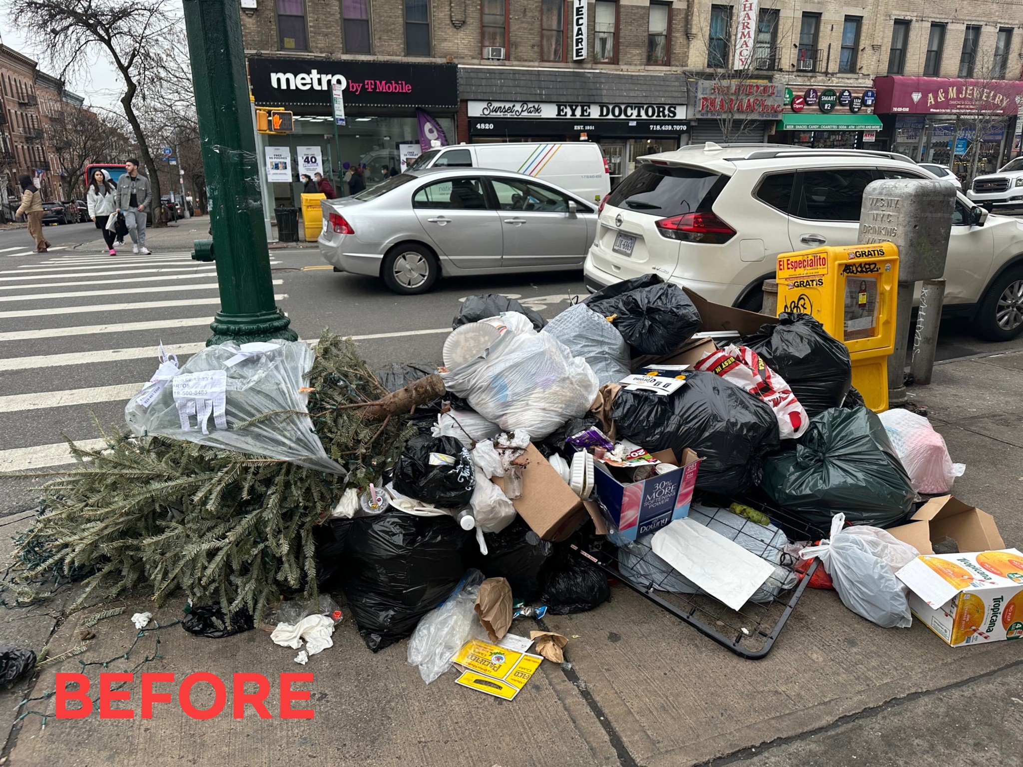 A pile of garbage on a city street corner, including bags, discarded boxes, and a christmas tree, with a 'before' sign at the bottom and traffic in the background.