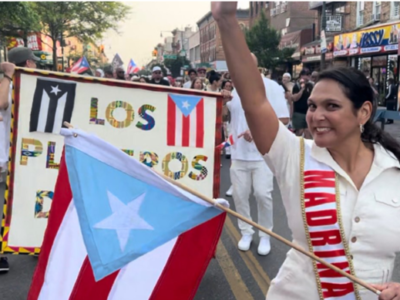 A woman in a white blouse and sash labeled "madrina" joyfully participates in a parade, waving a small puerto rican flag, next to a large decorated float with various cultural symbols and flags.