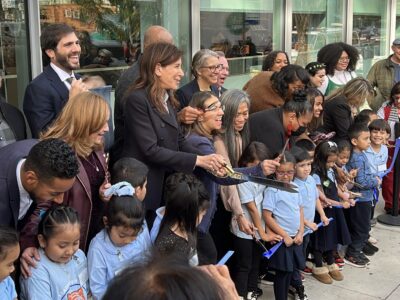 A diverse group of people, including children in school uniforms and adults, happily participates in a ribbon-cutting ceremony outdoors.