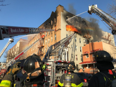 Firefighters using aerial ladders to hose down a blazing fire in a multi-story building while other firefighters watch from below. smoke and flames are visible.