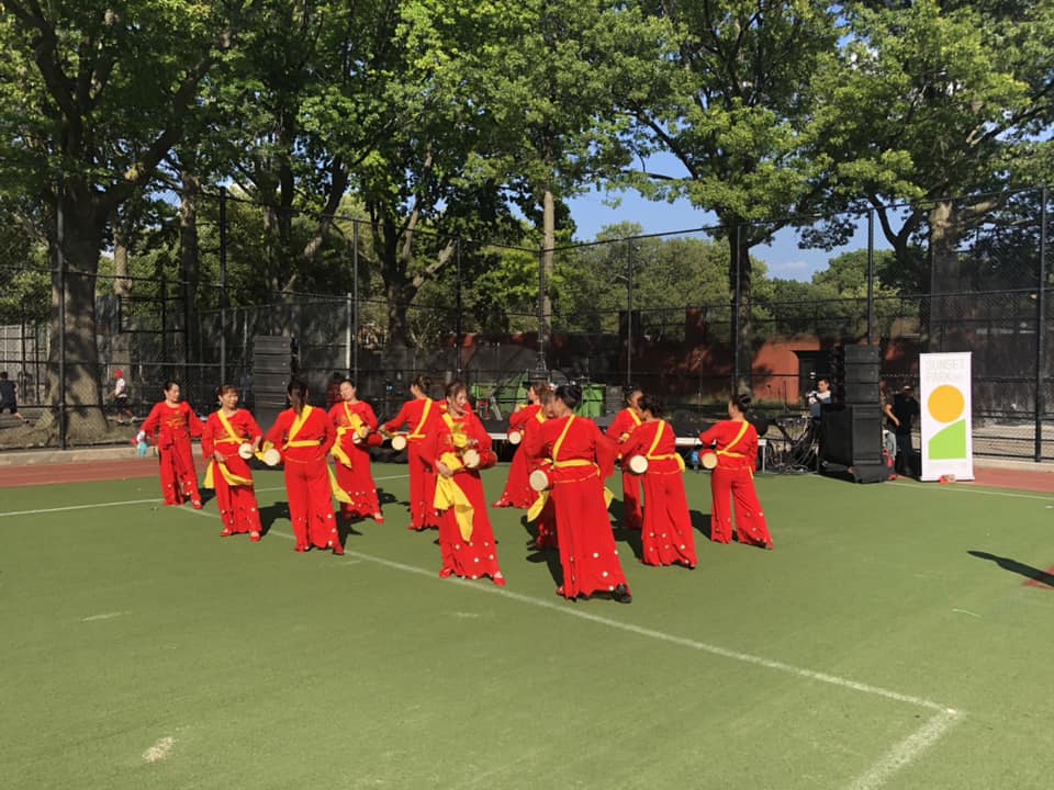 A group of women in red and yellow traditional dresses perform a dance with fans in an outdoor setting, with trees and a fence in the background.
