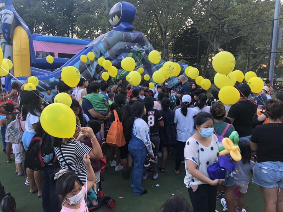 A crowded outdoor event with children and adults, many holding yellow balloons, near a large inflatable structure with a blue character on top.