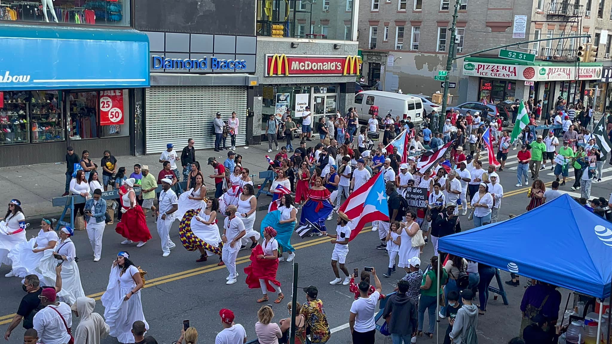 A vibrant street parade with people dressed in traditional and casual attire, waving puerto rican flags, dancing and marching down a city street lined with shops and fast food outlets.