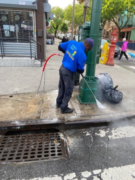 A worker in a blue vest uses a high-pressure water hose to clean a city sidewalk next to a green pole and black garbage bag.