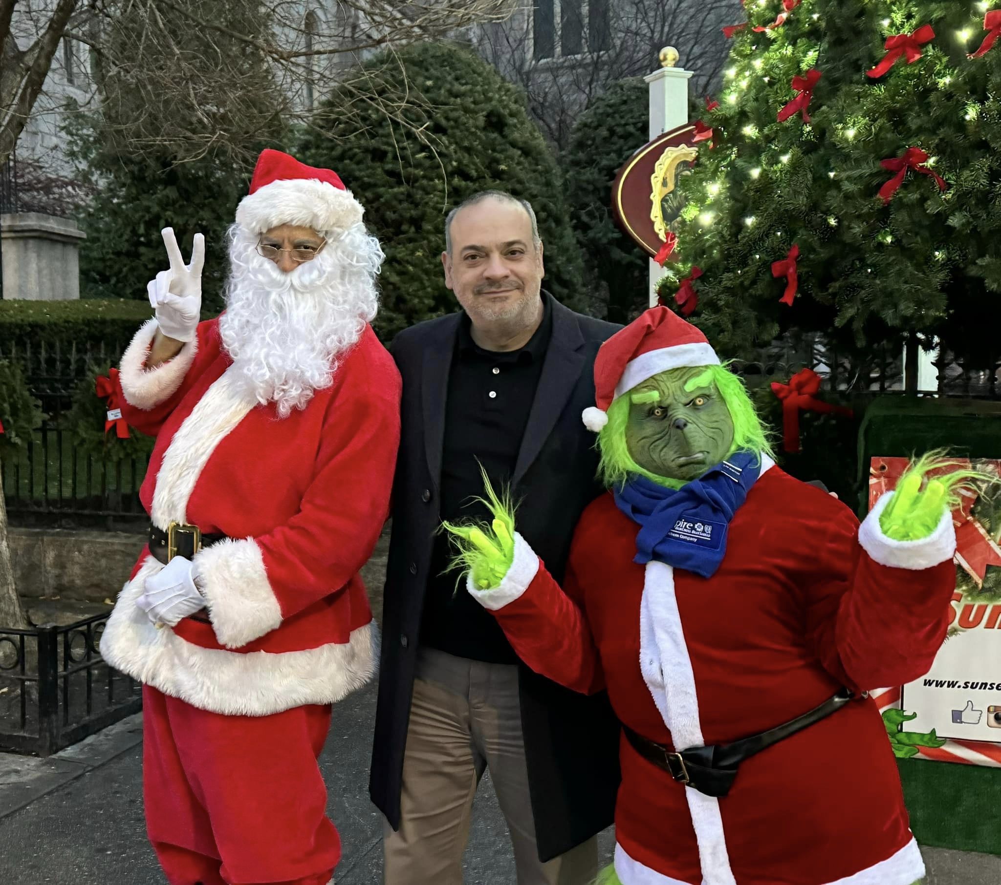 A man in a black jacket stands smiling between someone dressed as santa claus and another person costumed as the grinch, in front of a christmas tree.