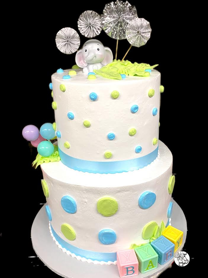 A two-tiered white cake adorned with colorful polka dots, featuring a top decoration of an elephant figurine and silver paper fans, surrounded by pastel-colored balloons and toy blocks spelling "baby.